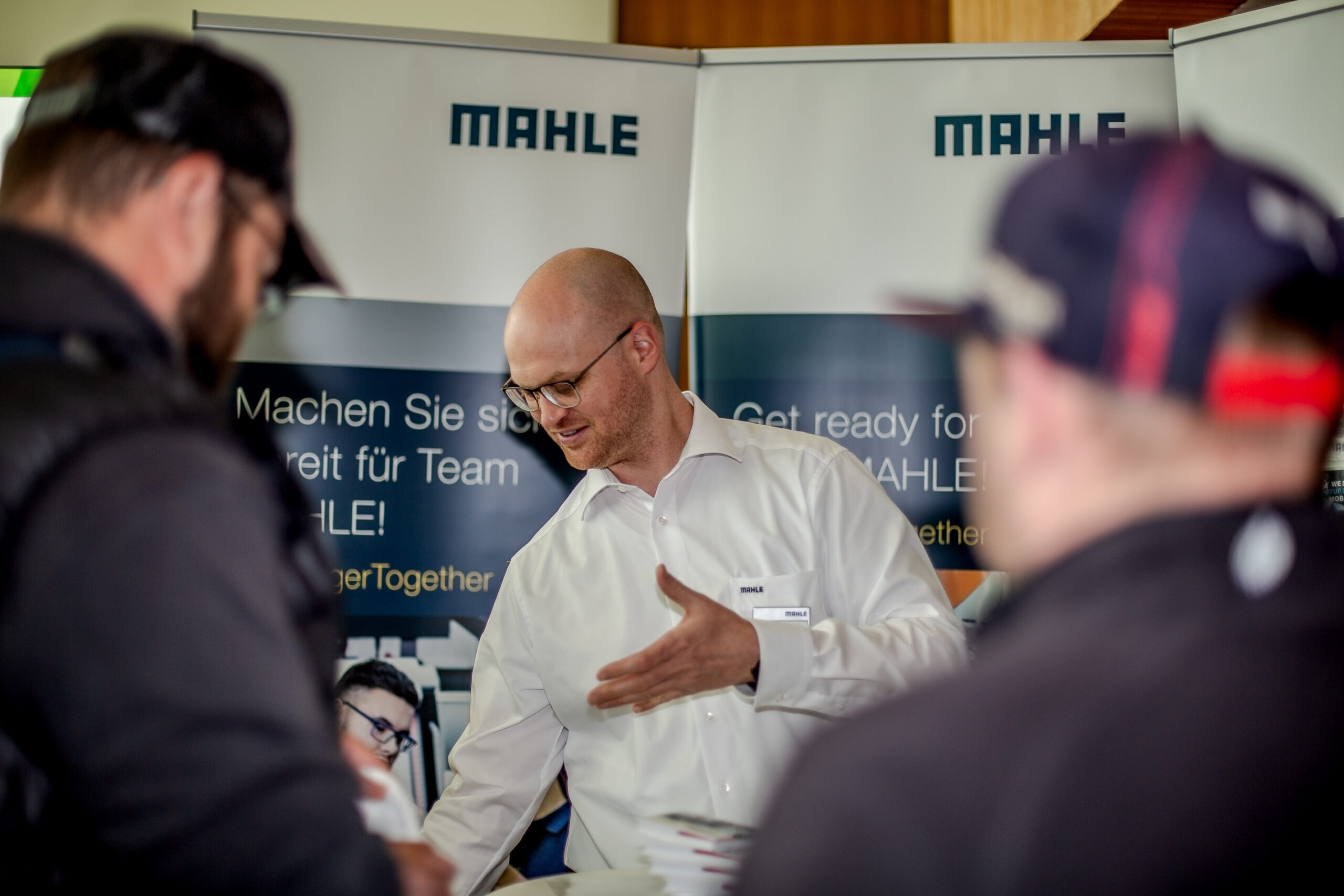 Ein Mann im weißen Hemd spricht mit zwei Personen an einem Stand, auf dem die Marke „MAHLE“ auf Bannern prominent zur Schau gestellt ist. Es sieht aus wie eine Indoor-Veranstaltung oder Messe, aber die Schilder in der Nähe deuten auf Kooperationen mit Institutionen wie der Hochschule Coburg hin und heben Innovationen und Partnerschaften hervor.