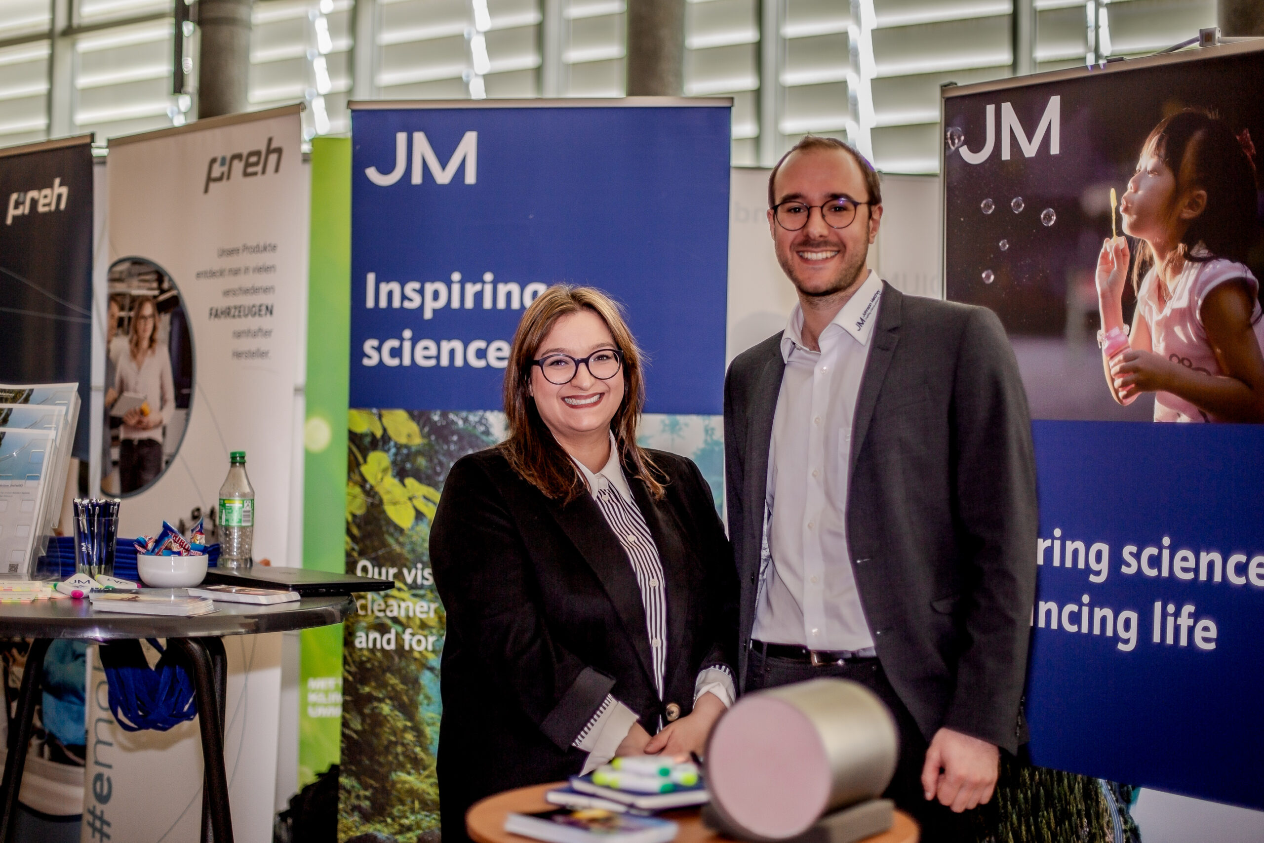 Zwei lächelnde Menschen bei einer wissenschaftlichen Veranstaltung der Hochschule Coburg. Sie stehen vor Bannern mit inspirierenden wissenschaftlichen Botschaften. Im Vordergrund ist ein Tisch mit Werbematerialien zu sehen, der den Innovationsgeist der Hochschule Coburg einfängt.