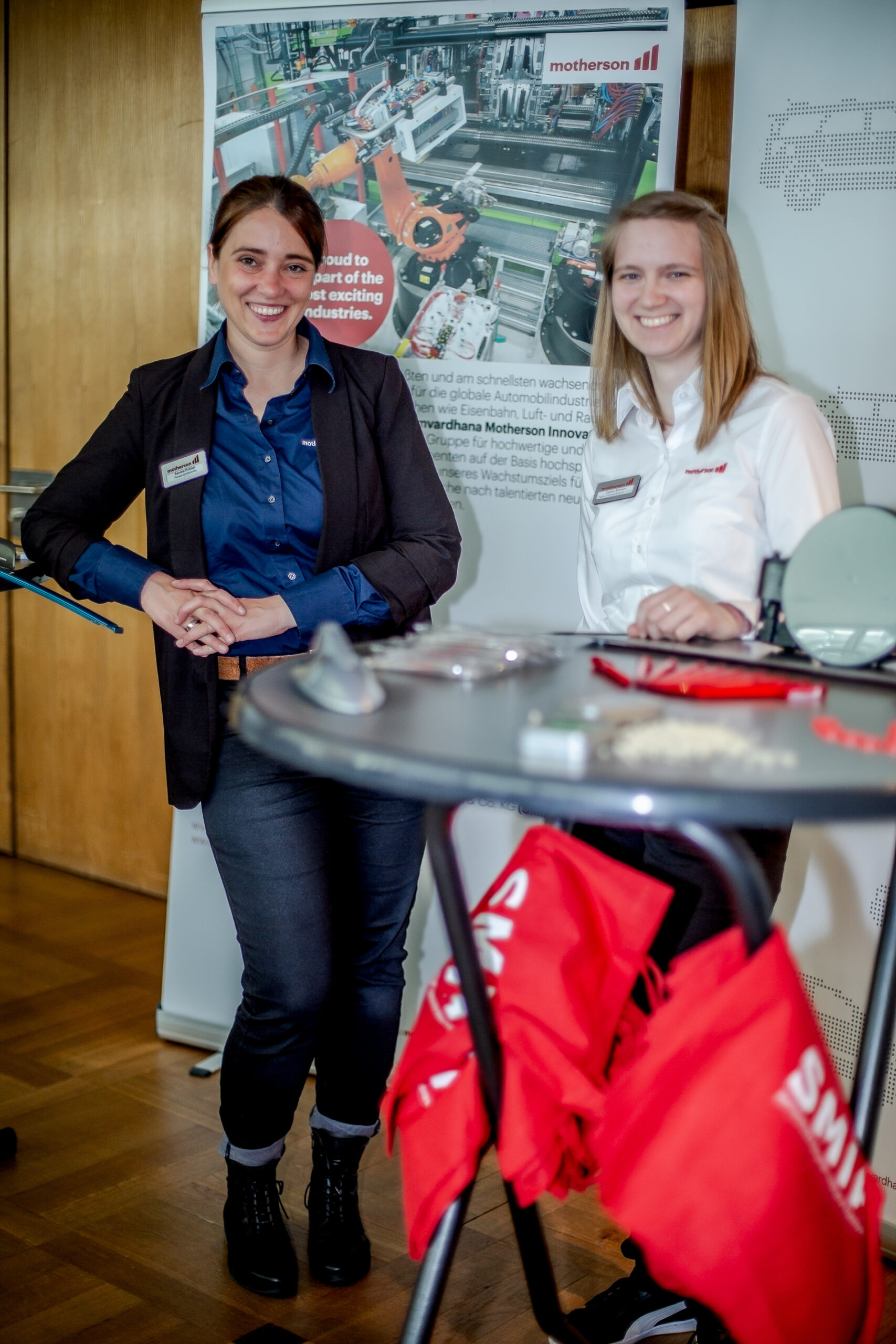 Zwei Frauen stehen lächelnd am Messestand der Hochschule Coburg. Die eine trägt einen schwarzen Blazer und ein blaues Hemd, die andere ein weißes. Auf dem Tisch sind rote Taschen und Gadgets ausgestellt, im Hintergrund ein Banner mit Industriebildern und Text.