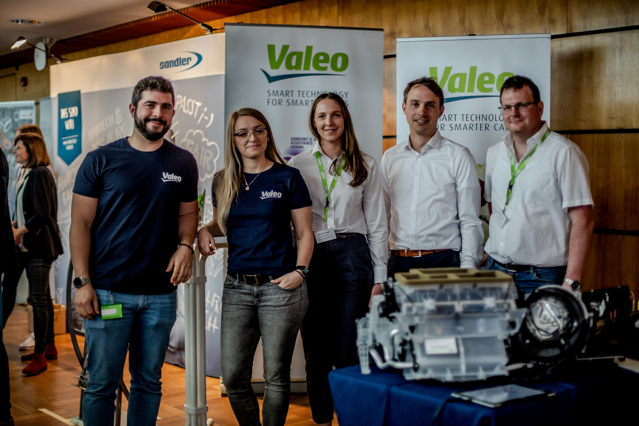 Eine Gruppe von fünf Personen der Hochschule Coburg steht zusammen an einem Messestand von Valeo, umgeben von Technologiedisplays und Werbebannern. Sie lächeln und sind in Freizeit- und Businesskleidung gekleidet.