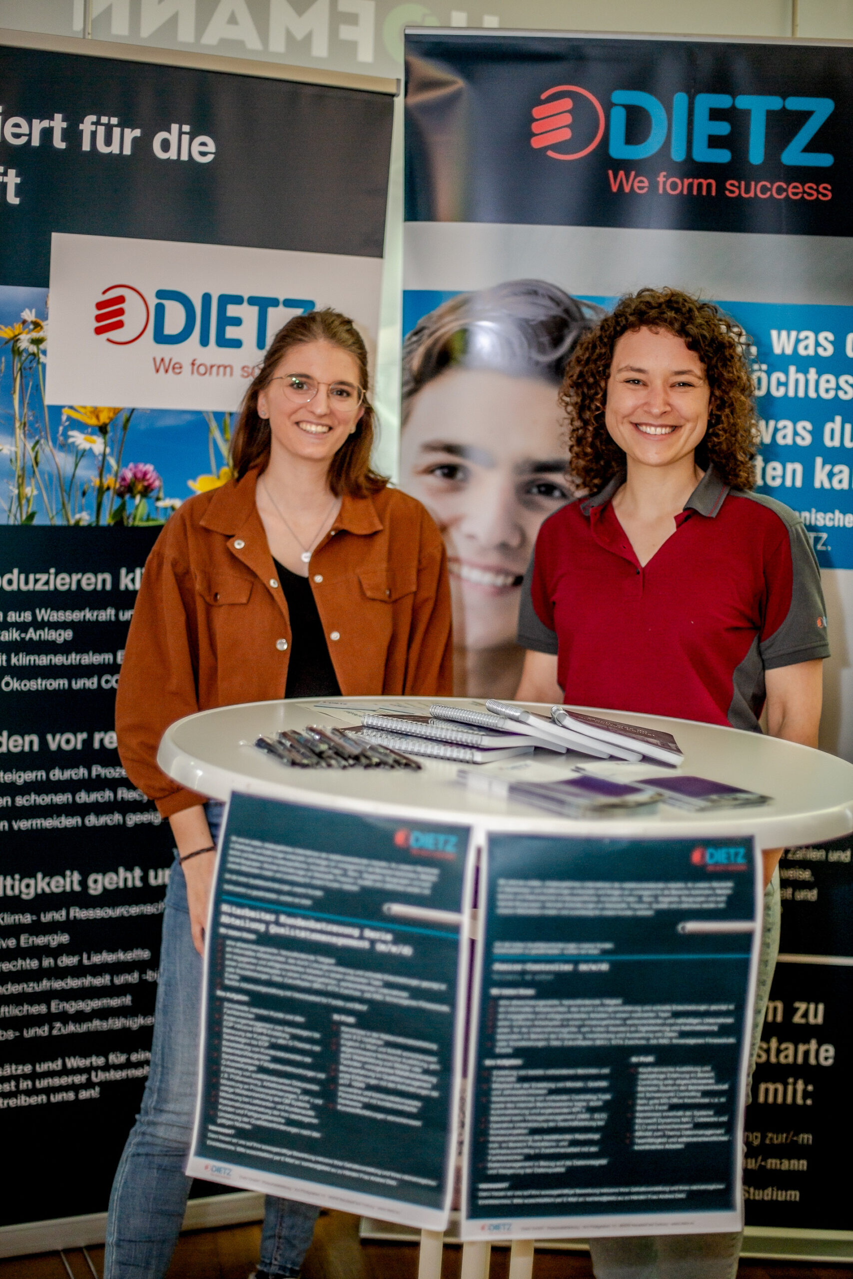 Zwei Personen lächeln an einem Stand mit „DIETZ Wir gestalten Erfolg“-Bannern und repräsentieren die Hochschule Coburg. Sie stehen hinter einem runden Tisch mit Broschüren und sorgen so für einen ansprechenden Auftritt auf der Veranstaltung oder Messe.