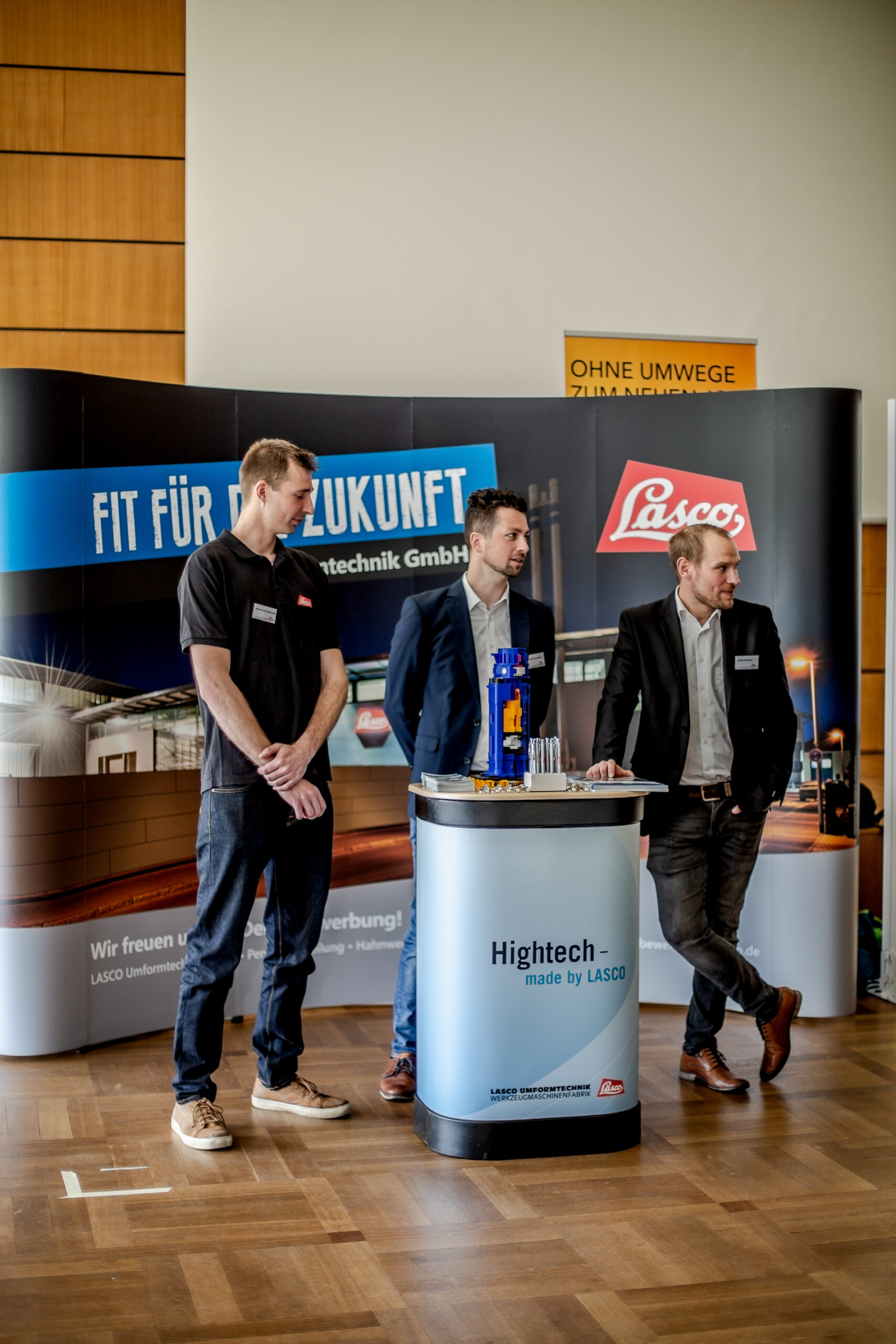 Drei Männer stehen in Business-Kleidung neben einem Stand mit den Aufschriften „Fit für die Zukunft“ und „Lasco“. Der Stand präsentiert hochtechnologische Produkte, die möglicherweise mit der Hochschule Coburg in Verbindung stehen. Holzverkleidung und ein Schild darüber vervollständigen das professionelle Ambiente.