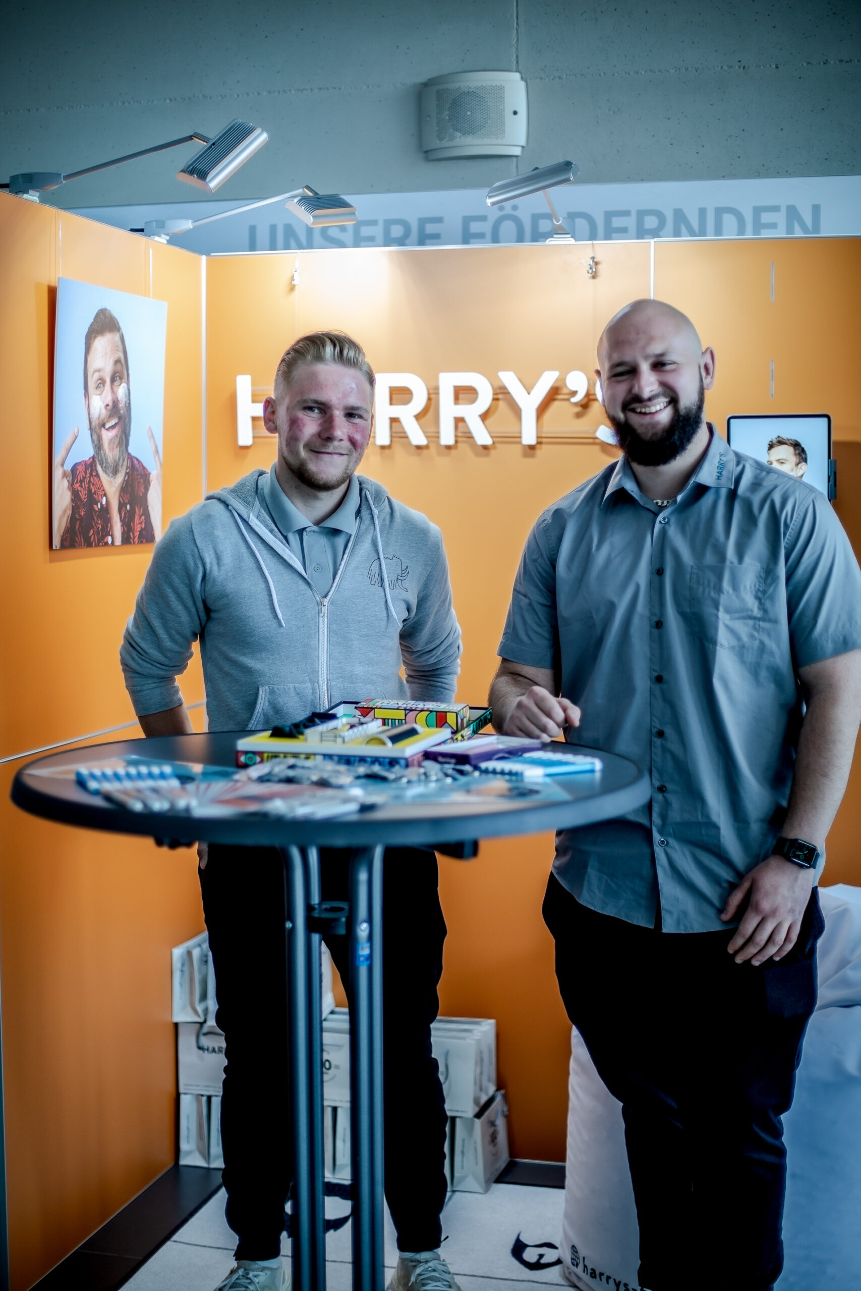 Zwei Männer stehen und lächeln an einem Werbestand für „Harry’s“ auf der Veranstaltung der Hochschule Coburg. Im Hintergrund sind Branding und Fotos zu sehen. Jeder von ihnen hält ein Produkt in der Hand, und auf dem runden Tisch vor ihnen sind verschiedene Artikel ausgestellt.