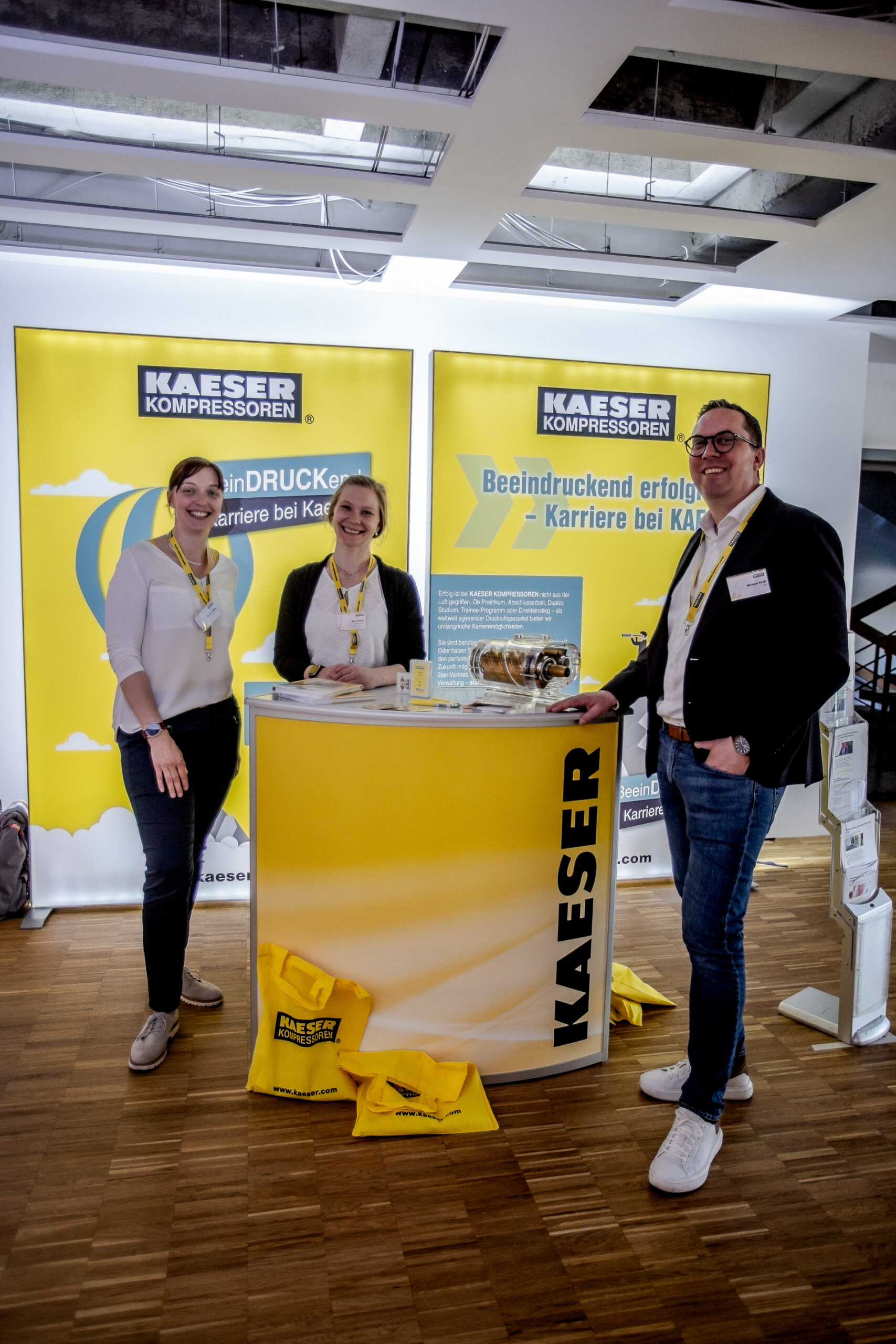 Drei Personen stehen lächelnd um einen gelben Stand mit dem Logo „KAESER Kompressoren“ herum und scheinen an einer Veranstaltung der Hochschule Coburg teilzunehmen. Vor dem Stand stehen gelbe Taschen mit dem gleichen Logo. Der Stand ist in einem Innenbereich mit Holzboden und heller Beleuchtung.