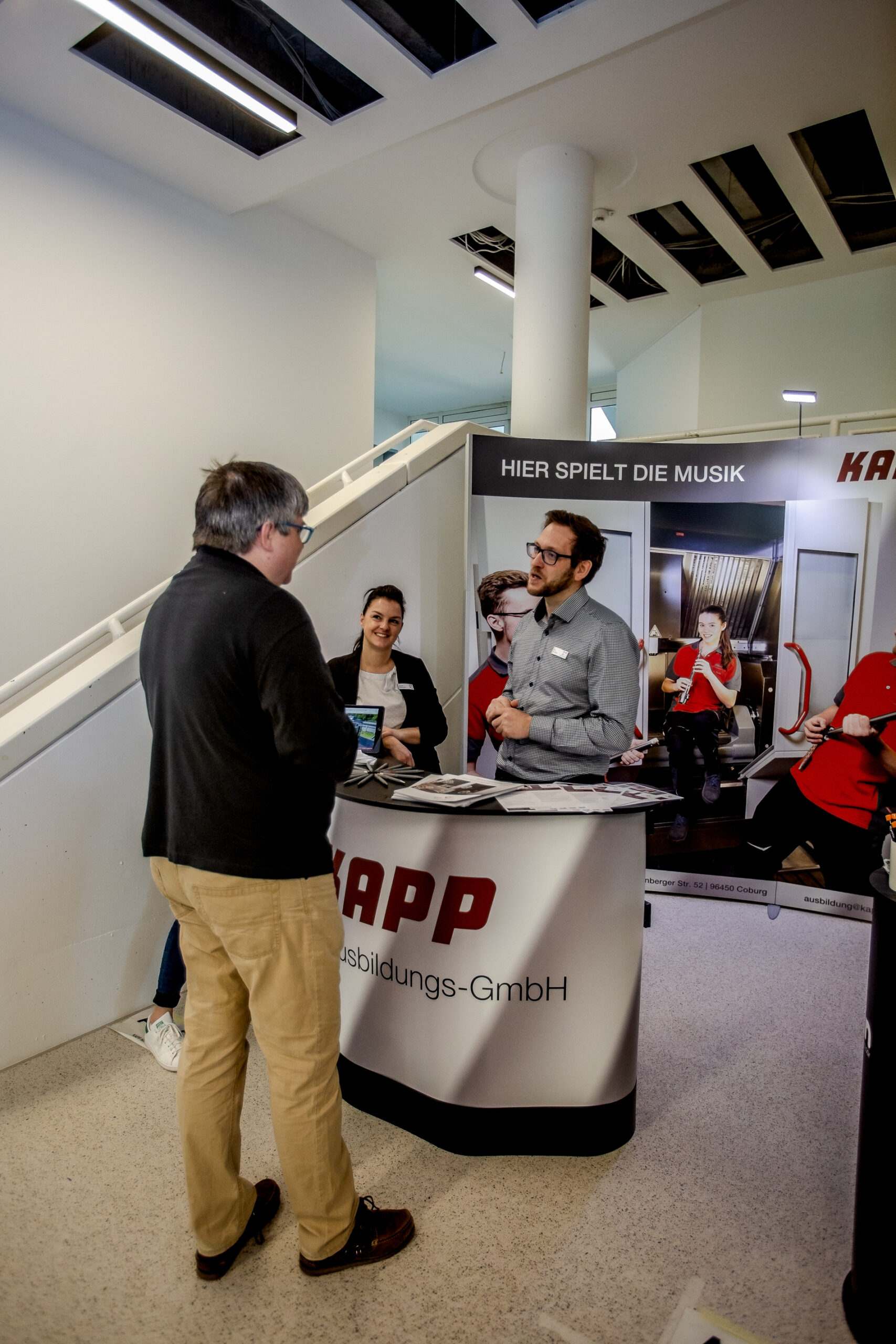 Ein Mann in dunkler Jacke unterhält sich während einer Veranstaltung an der Hochschule Coburg mit zwei Personen an einem Stand mit der Aufschrift „KAPP“. Der Stand zeigt im Hintergrund Bilder und befindet sich in einer eleganten, modernen Innenumgebung.