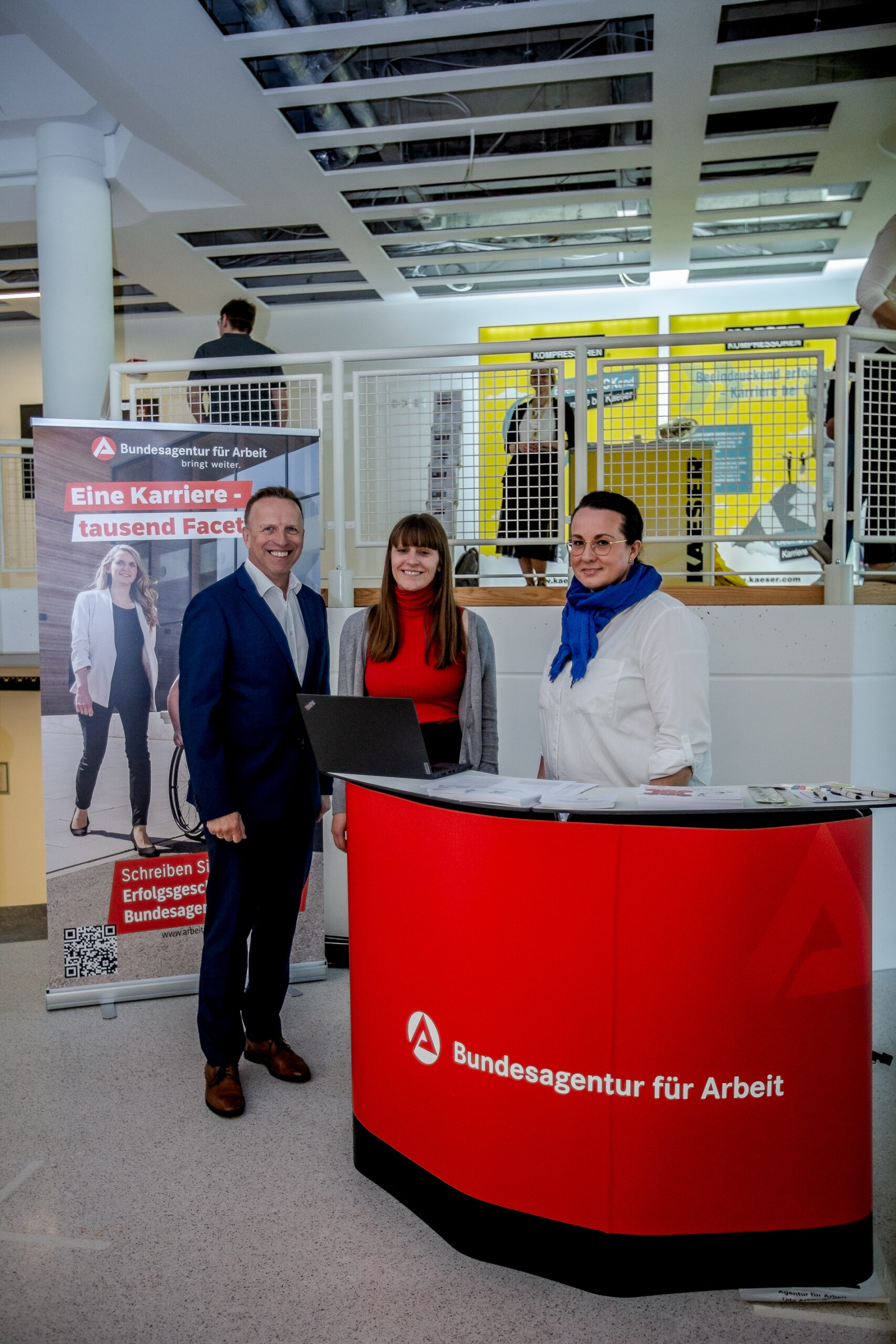 Drei Personen stehen hinter einem Schreibtisch mit dem Logo der Bundesagentur für Arbeit. Zwei Frauen und ein Mann, Vertreter der Hochschule Coburg, posieren neben einem Laptop und einem Werbebanner. Die Umgebung ist hell und modern, im Hintergrund sind eine Treppe und ein offenes Geländer zu sehen.