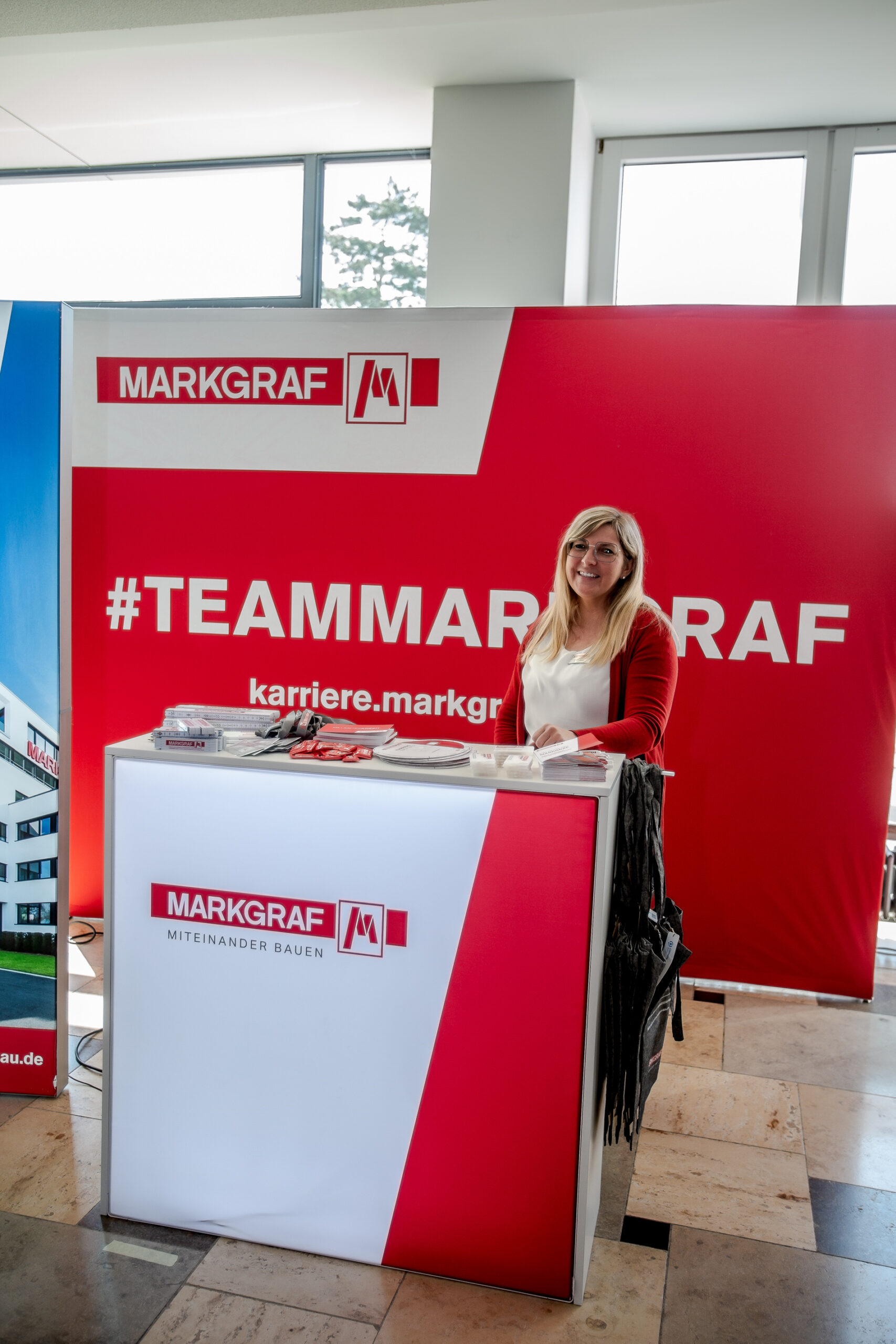 Eine Frau steht an einem weißen Promotionstand, der mit rot-weißem Markgraf-Branding geschmückt ist und den Slogan „#TEAMMARKGRAF“ präsentiert. Sie lächelt und hält Papiere in der Hand, im Hintergrund sind große Banner zu sehen. Der Stand zeigt stolz die Zusammenarbeit mit der Hochschule Coburg.