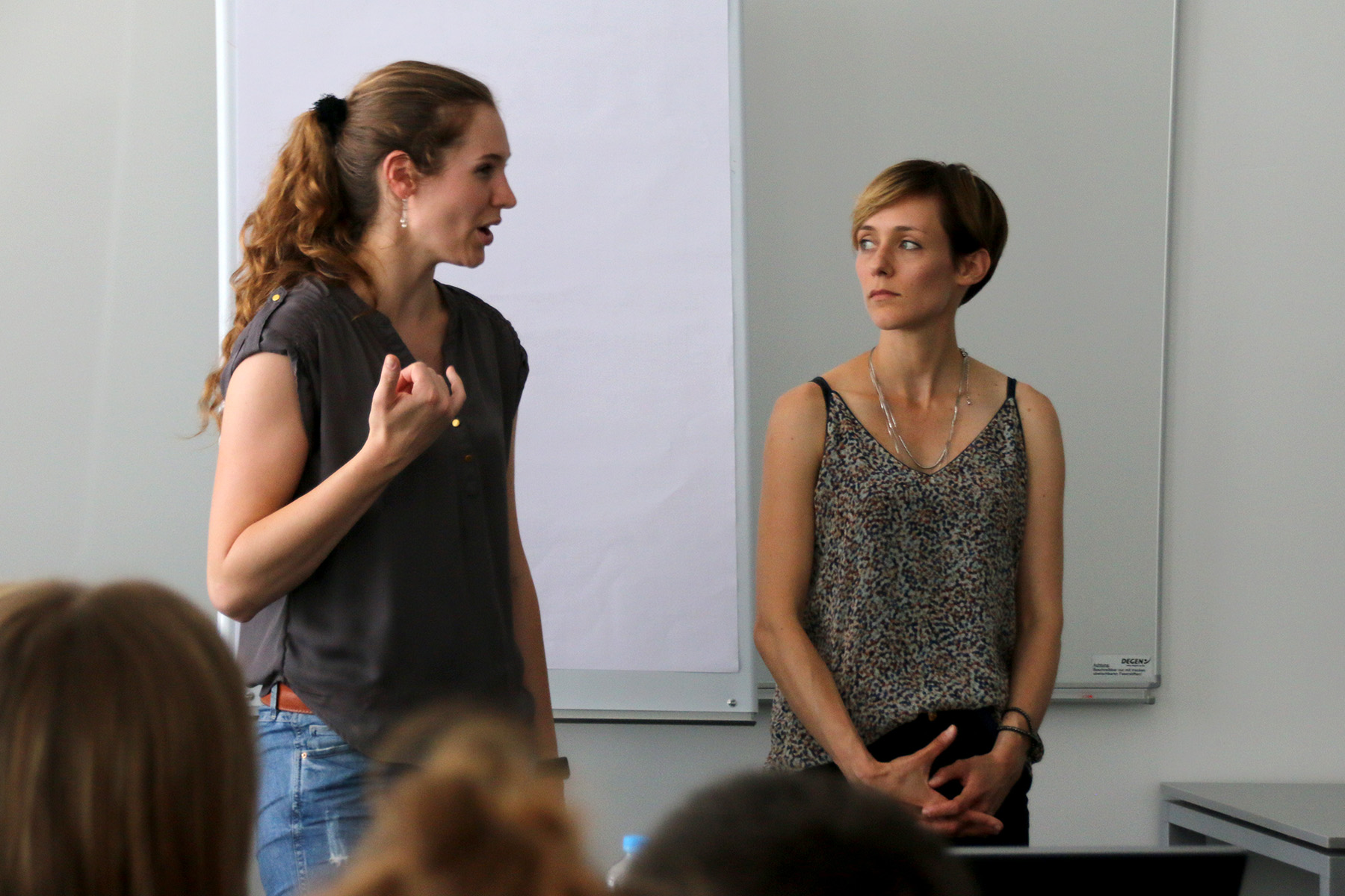 Zwei Frauen stehen vor einem leeren Flipchart an der Hochschule Coburg. Die Frau links spricht und gestikuliert, während die Frau rechts mit gefalteten Händen zuhört. Im Vordergrund ist ein unscharfes Publikum zu sehen, das von ihrer spannenden Präsentation gefesselt ist.