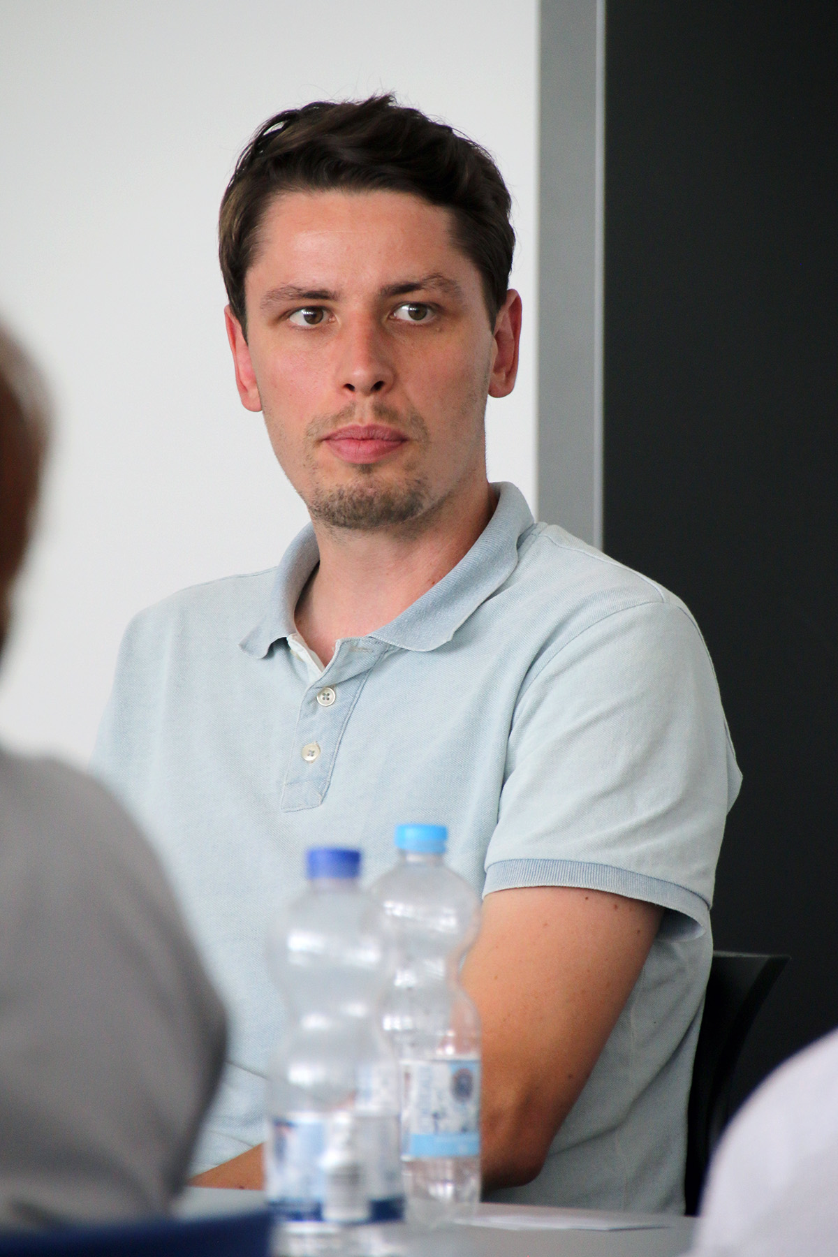 Ein Mann in einem hellblauen Poloshirt sitzt an einem Tisch, vor ihm stehen Wasserflaschen, und blickt zur Seite. Der Hintergrund, der an die Hochschule Coburg erinnert, ist eine schlichte, verschwommene Innenkulisse.