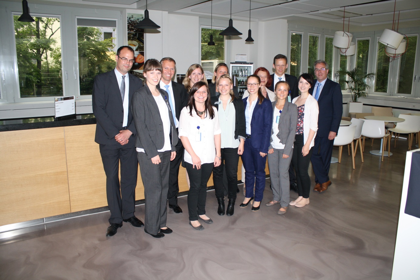 Eine Gruppe von zwölf Berufstätigen in Business-Kleidung steht lächelnd in einem Büroküchenbereich der Hochschule Coburg. Große Fenster, Tische und Stühle bilden den Hintergrund und schaffen eine helle und einladende Atmosphäre.