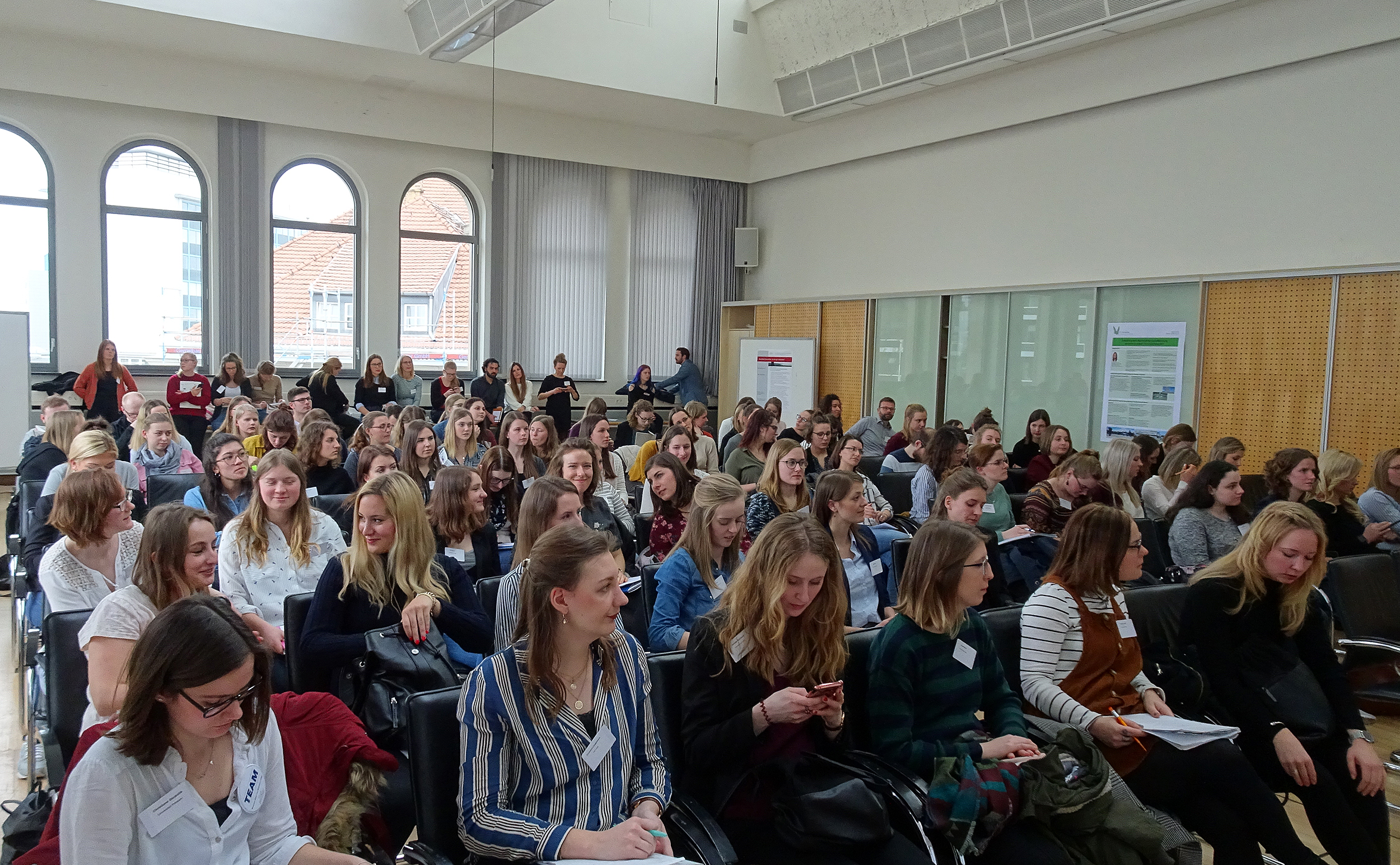 Eine große Gruppe von Menschen, überwiegend Frauen, sitzt in einem gut beleuchteten Konferenzraum der Hochschule Coburg. Sie sind konzentriert und scheinen einer Präsentation zuzuhören. Einige halten Notizblöcke in den Händen, an den Wänden hängen Plakate. Große Fenster im Hintergrund lassen reichlich Tageslicht herein.