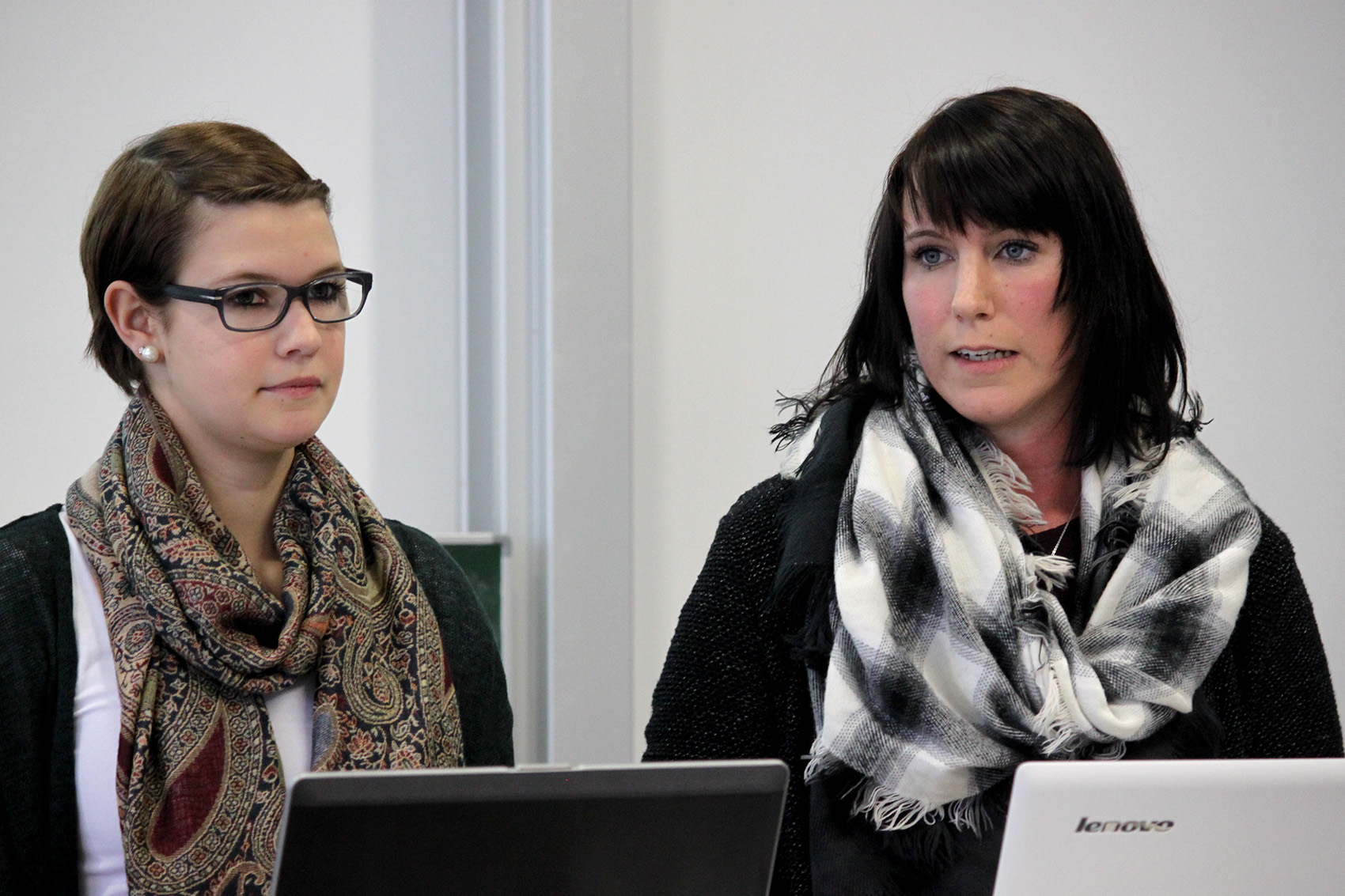 Zwei Frauen, beide mit Kopftuch und eine mit Brille, stehen in einer Präsentation an der Hochschule Coburg. Sie sitzen neben Laptops vor einem Whiteboard und sind voll auf ihre Aufgabe konzentriert.