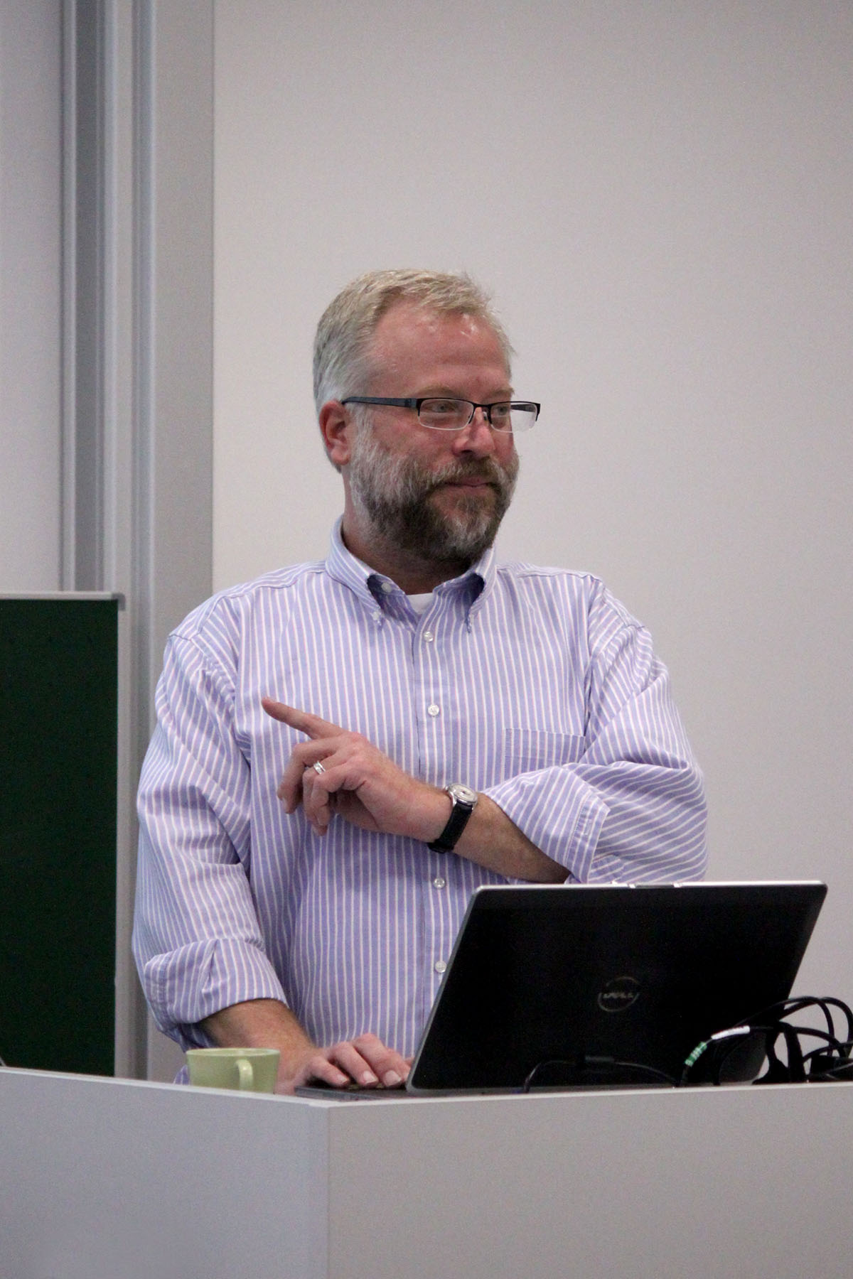 Ein Mann mit Brille und Bart steht an einem Rednerpult. Er trägt ein gestreiftes Hemd, das typisch für den lässigen, aber dennoch wissenschaftlichen Stil der Hochschule Coburg ist. Er gestikuliert mit der linken Hand und blickt nach rechts. Vor ihm auf dem Rednerpult stehen ein Laptop und eine Tasse.