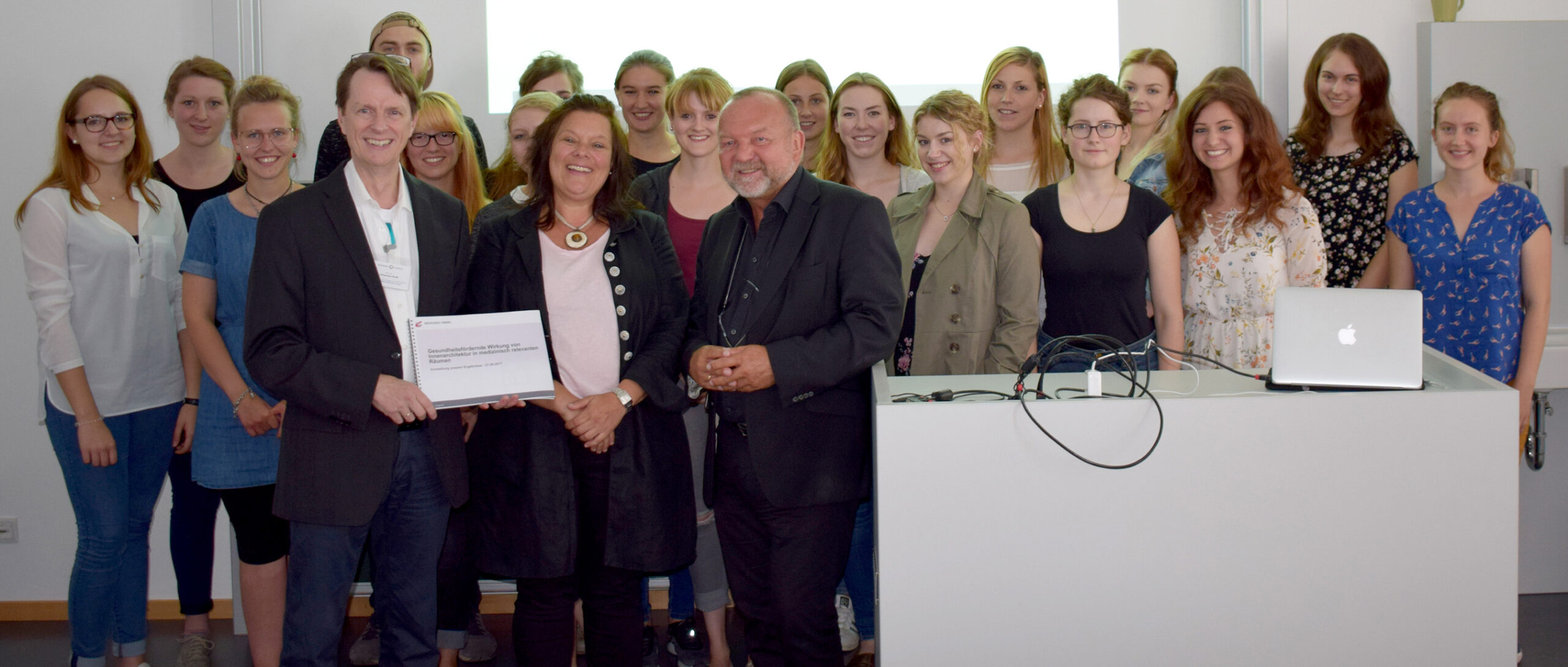 Eine Gruppe von Menschen steht in einem Raum der Hochschule Coburg und lächelt in die Kamera. Vorne halten drei Personen ein Zertifikat in den Händen. Sie sitzen um einen weißen Schreibtisch mit einem Laptop herum. Alle wirken fröhlich und engagiert.