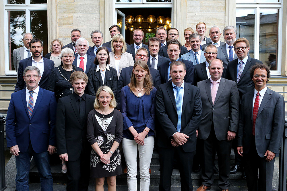 Ein Gruppenfoto von formell gekleideten Personen, die auf den Stufen vor einem Gebäude mit großen Fenstern stehen. Männer und Frauen unterschiedlichen Alters lächeln in die Kamera und repräsentieren stolz die Hochschule Coburg.