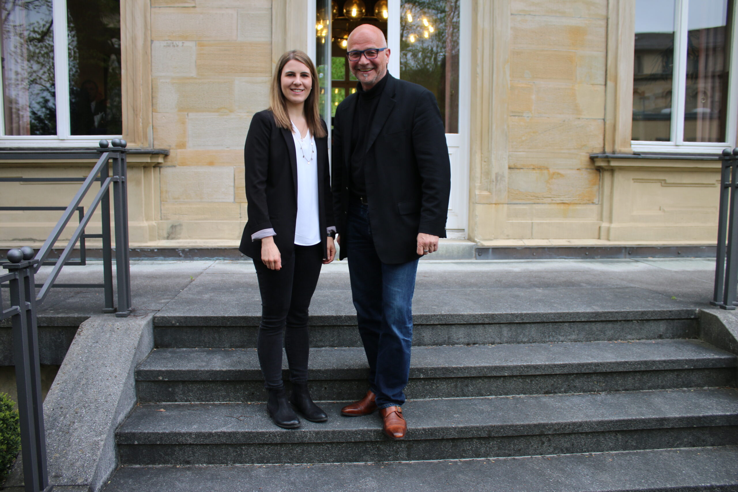 Ein Mann und eine Frau stehen auf den Stufen vor der Hochschule Coburg. Beide tragen schwarze Jacken. Die Frau trägt ein weißes Hemd und eine schwarze Hose, während der Mann blaue Jeans und braune Schuhe trägt. Sie lächeln herzlich in die Kamera und fangen einen Moment der Verbundenheit auf dem Campus ein.