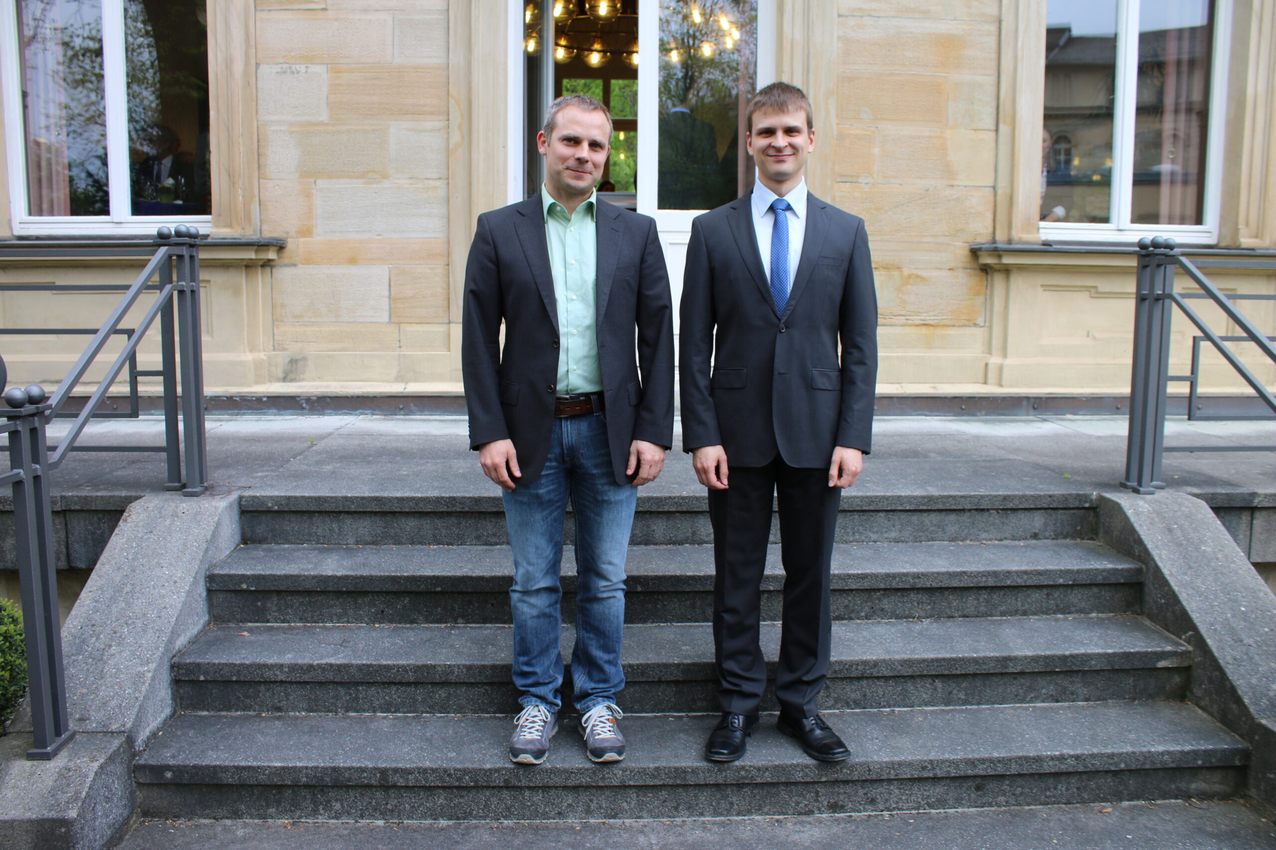 Zwei Männer stehen auf Steinstufen vor einem Gebäude mit großen Fenstern der Hochschule Coburg. Der Mann links trägt ein legeres Outfit mit grünem Hemd und Jeans, während der Mann rechts einen formellen Anzug und Krawatte trägt. Beide lächeln.