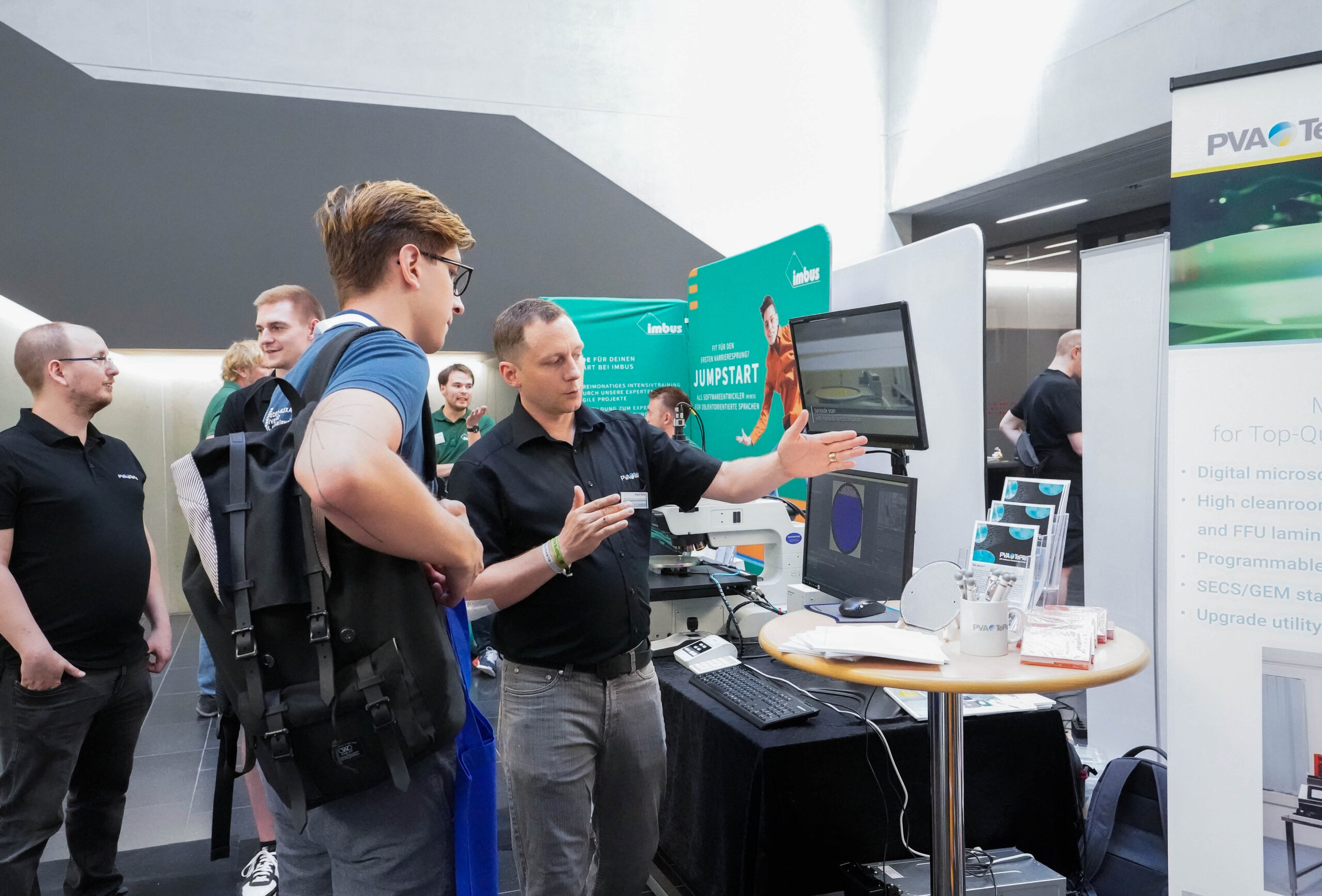 Ein Mann mit Rucksack hört während einer Veranstaltung einem Verkäufer am Technikstand der Hochschule Coburg zu. Sie stehen an einem Tisch mit elektronischen Geräten und Bildschirmen. Im Hintergrund sind mehrere andere Teilnehmer und Stände zu sehen, die zur geschäftigen Atmosphäre beitragen.