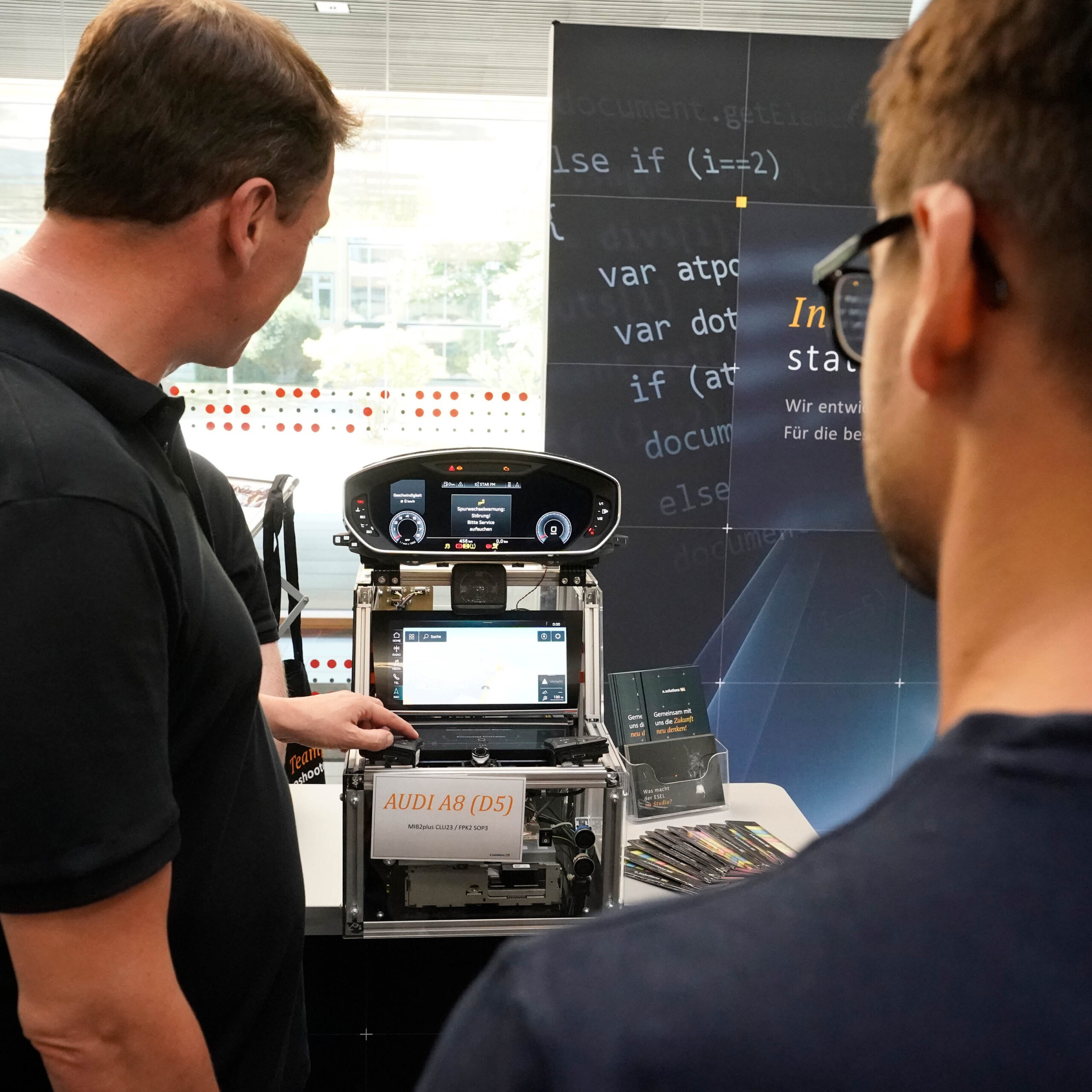 Zwei Personen der Hochschule Coburg beobachten auf einem Ausstellungsständer ein elektronisches Setup mit der Aufschrift „AUDI A8 (D5)“. Zu sehen sind verschiedene Bildschirme und Komponenten, auf den Hintergrundtafeln ist teilweise Text verdeckt, was ihre Neugier und ihr akademisches Interesse weckt.