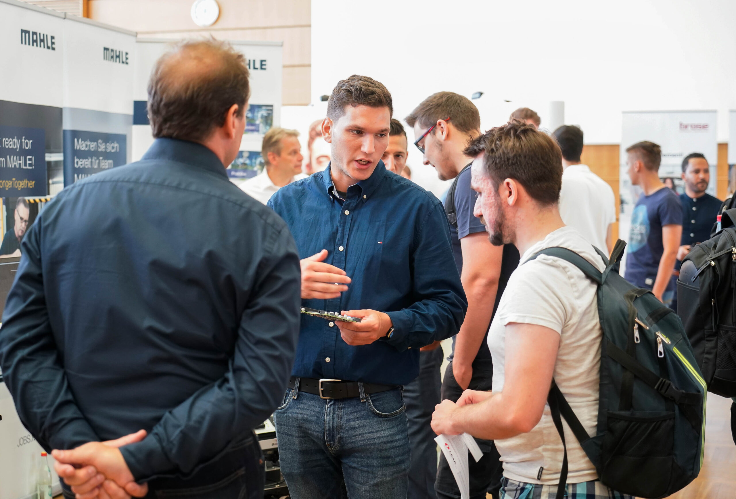 Eine Gruppe von Männern unterhält sich bei einer Indoor-Networking-Veranstaltung der Hochschule Coburg. Ein Mann im blauen Hemd spricht und gestikuliert dabei mit der Hand. Andere im Hintergrund unterhalten sich, umgeben von Informationsbannern und in professioneller Kleidung.