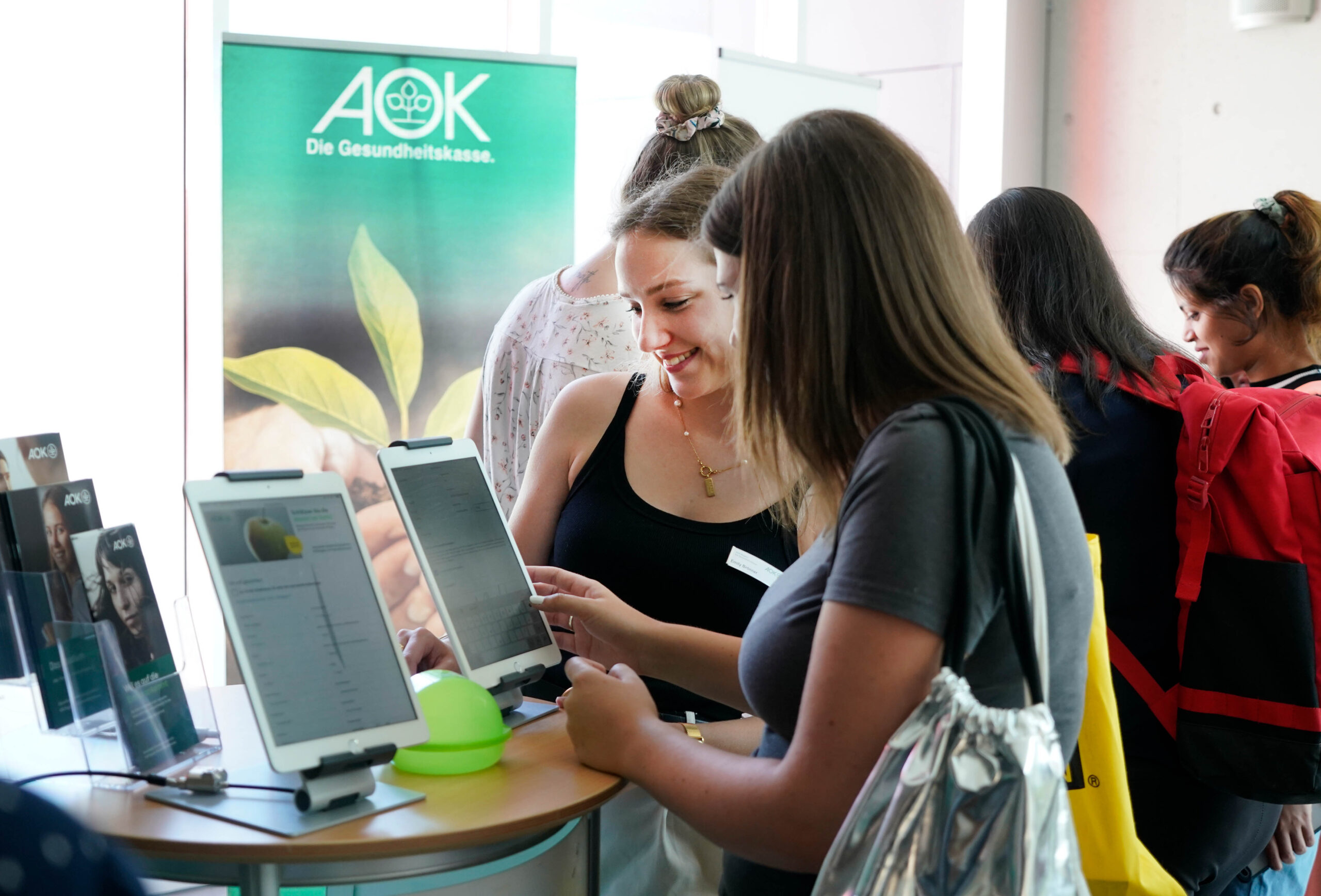 Zwei Frauen arbeiten an einem Stand mit einem AOK-Banner und umgeben von Broschüren mit Bezug zur Hochschule Coburg mit digitalen Tablets. Eine Frau lächelt, während die andere auf den Bildschirm schaut. Im Hintergrund tummeln sich weitere Menschen, die in strömendes Sonnenlicht getaucht sind.