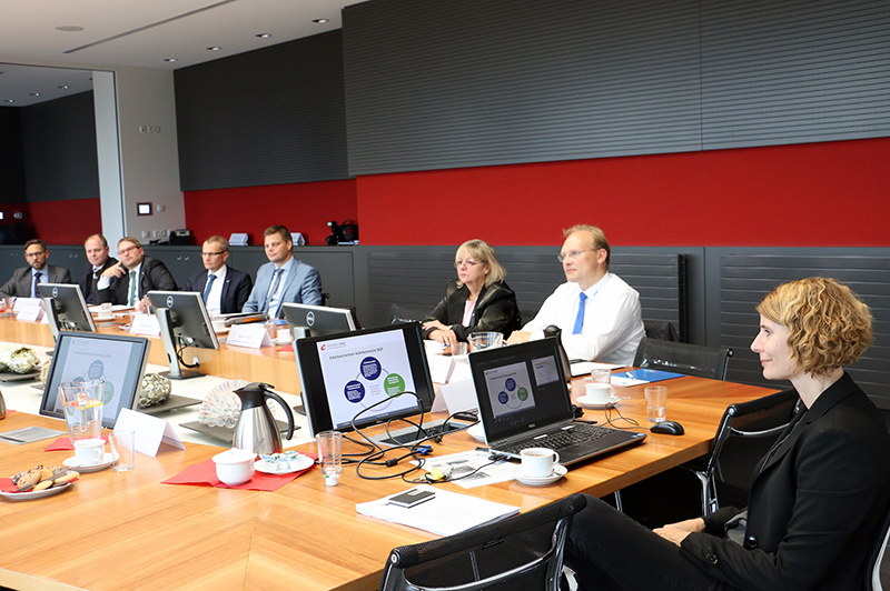 Eine Gruppe von Fachleuten in Business-Kleidung sitzt an einem großen hölzernen Konferenztisch in der Hochschule Coburg. Laptops und eine Projektionsleinwand zeigen grafische Präsentationen, während Kaffeetassen und Dokumente über den Tisch verstreut sind. Den Hintergrund bildet eine rote Wand.