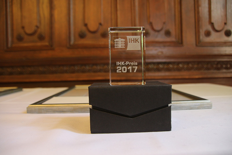 Eine Trophäe mit der Aufschrift „IHK-Preis 2017“ steht elegant auf einem schwarzen Kasten, der auf einer weißen Tischdecke ruht. Dahinter akzentuiert eine Holzvertäfelung die Szenerie und erinnert an eine Preisverleihung an der Hochschule Coburg.