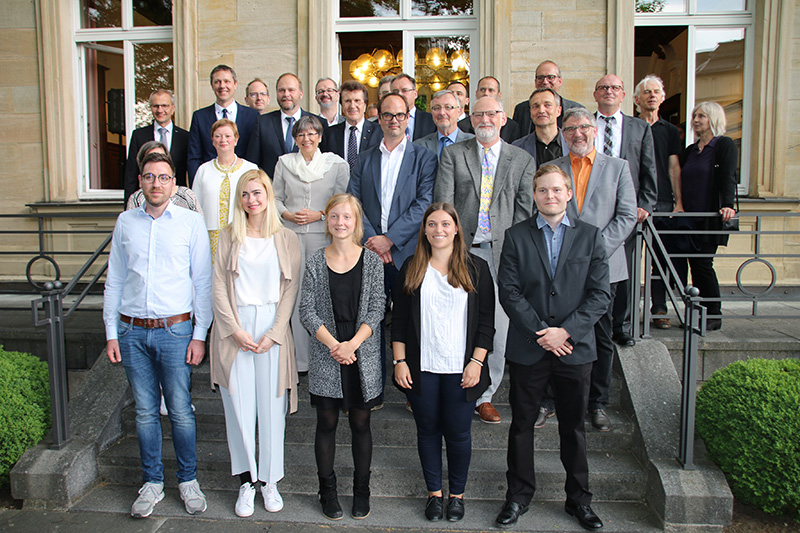 Ein Gruppenfoto zeigt Menschen, die auf den Stufen vor einem Gebäude stehen, möglicherweise der Hochschule Coburg. Die Männer und Frauen in formeller und halbformeller Kleidung posieren lächelnd zusammen, was auf eine Versammlung oder Veranstaltung im Freien schließen lässt.