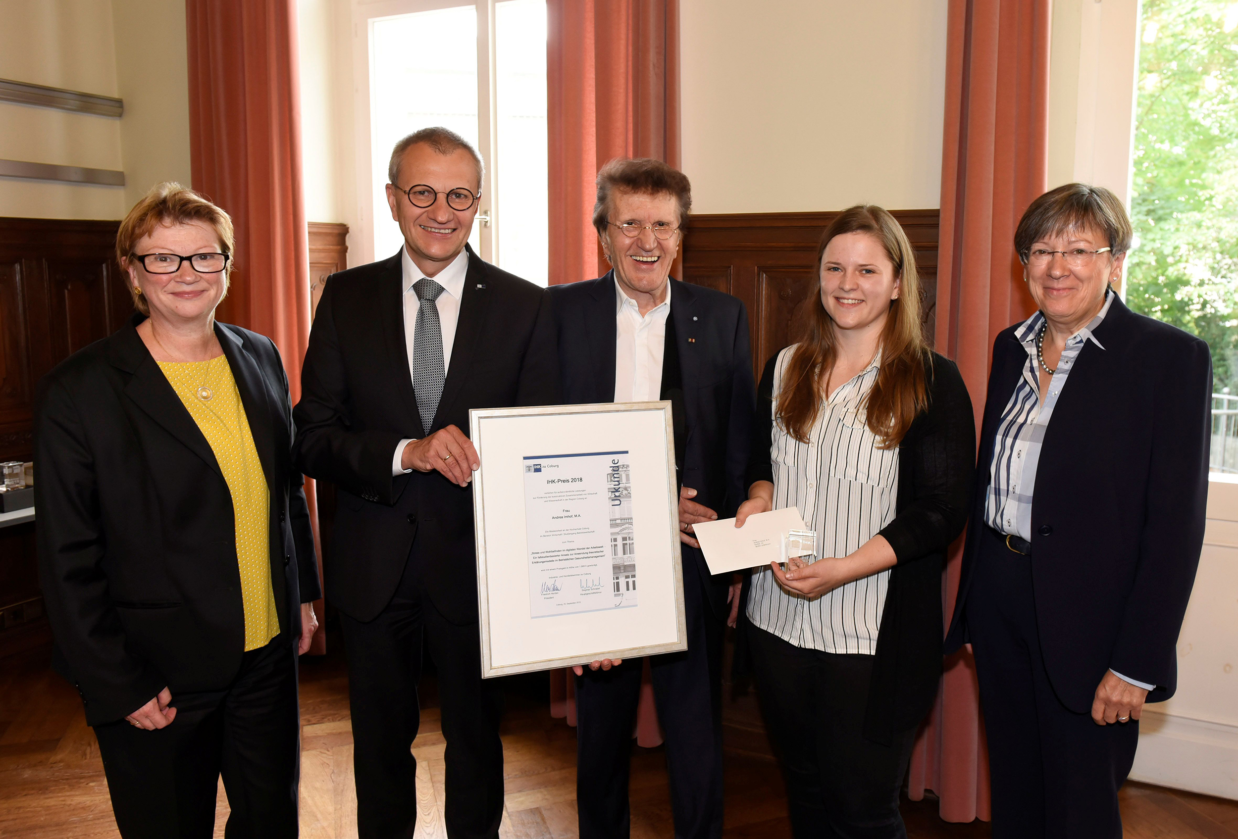 Fünf Menschen stehen zusammen im Raum und lächeln. Ein Mann im Anzug hält ein gerahmtes Zertifikat der Hochschule Coburg in der Hand. Eine junge Frau neben ihm hält einen weißen Umschlag. Sie befinden sich in einem Raum mit Holzvertäfelung und hohen Fenstern.