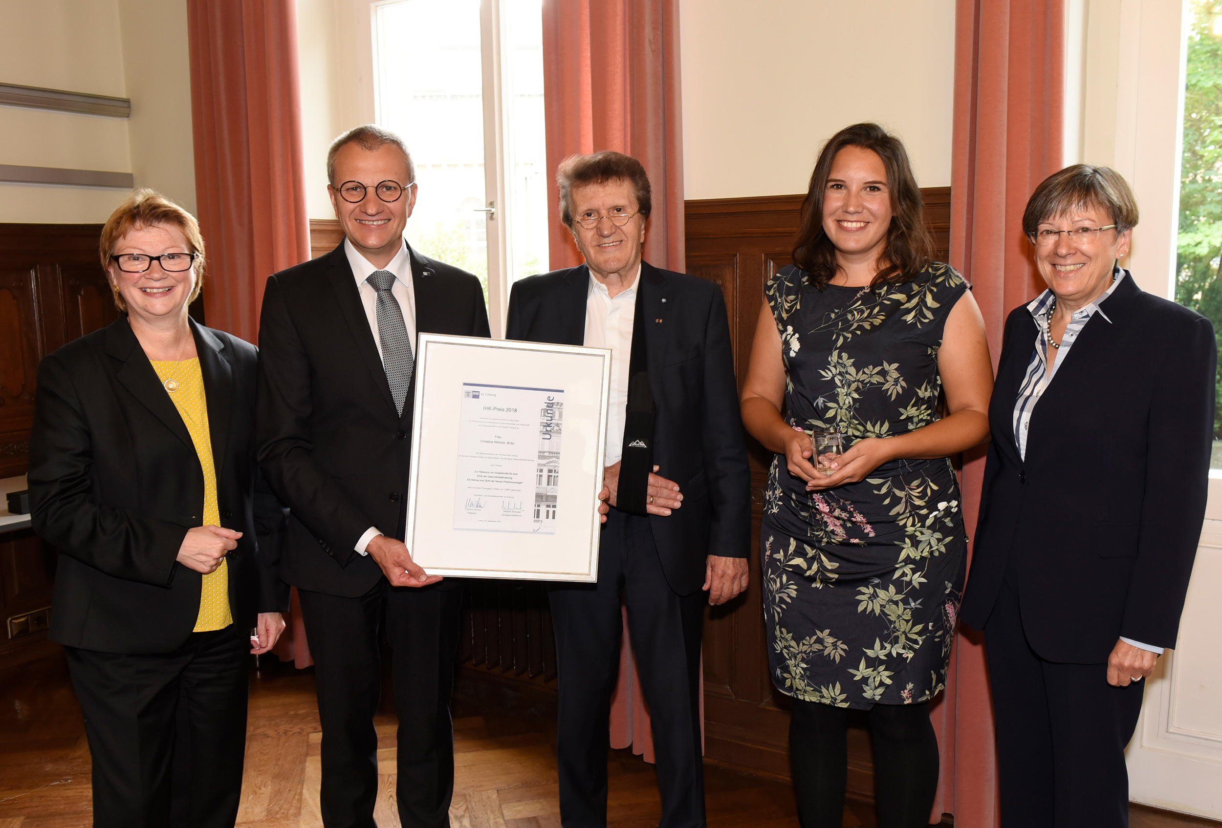 Fünf Menschen stehen zusammen in einem Raum mit roten Vorhängen an der Hochschule Coburg. Zwei Menschen in der Mitte halten eine große Urkunde in den Händen. Alle lächeln und sind in formeller Kleidung gekleidet. Die Gruppe scheint eine Leistung zu feiern.