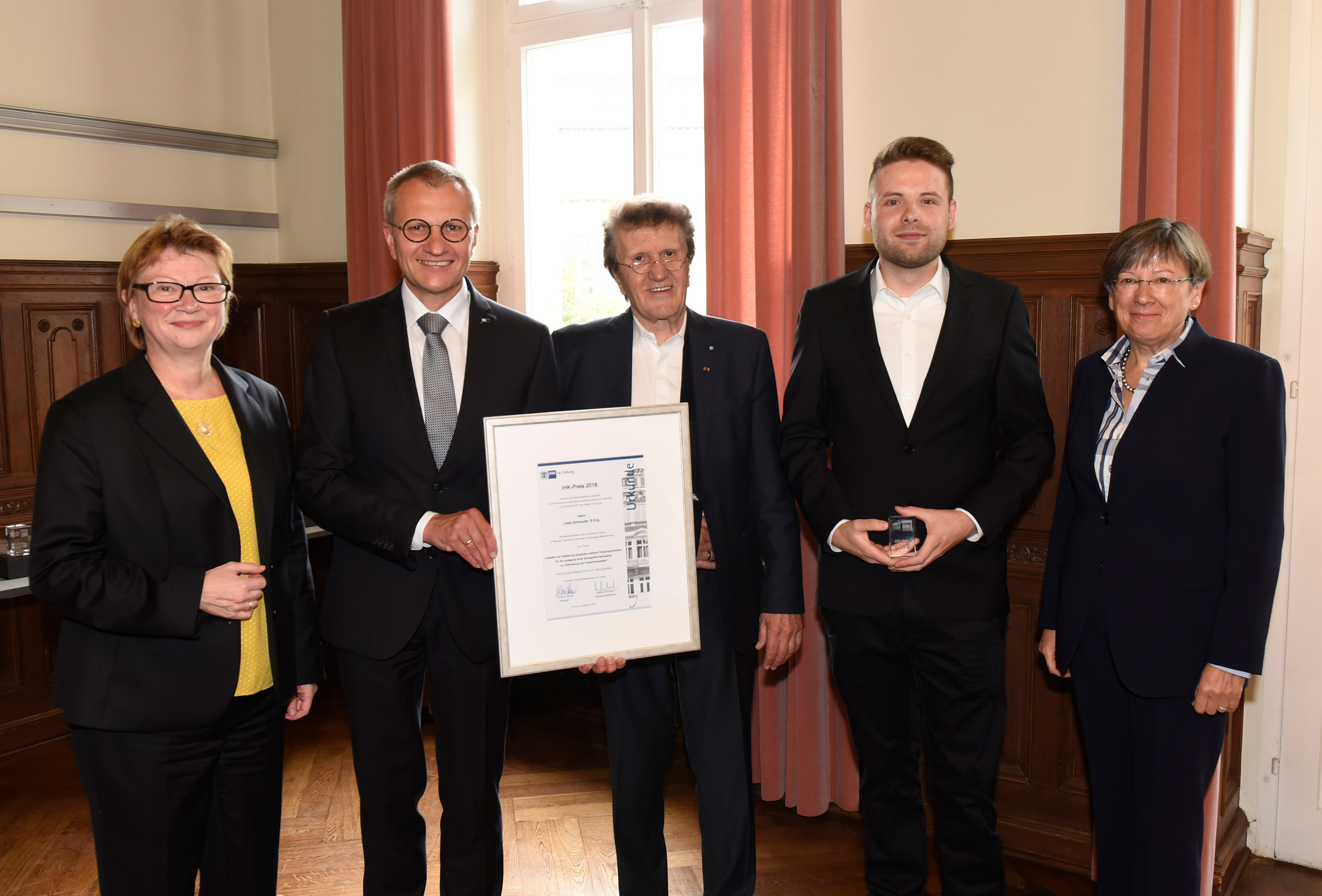 Eine Gruppe von fünf Personen in formeller Kleidung steht in einem Innenbereich der Hochschule Coburg zusammen. Eine Person hält ein gerahmtes Zertifikat. Sie lächeln und posieren für ein Foto in einem Raum mit Holzvertäfelung und einem mit Vorhängen geschmückten Fenster.