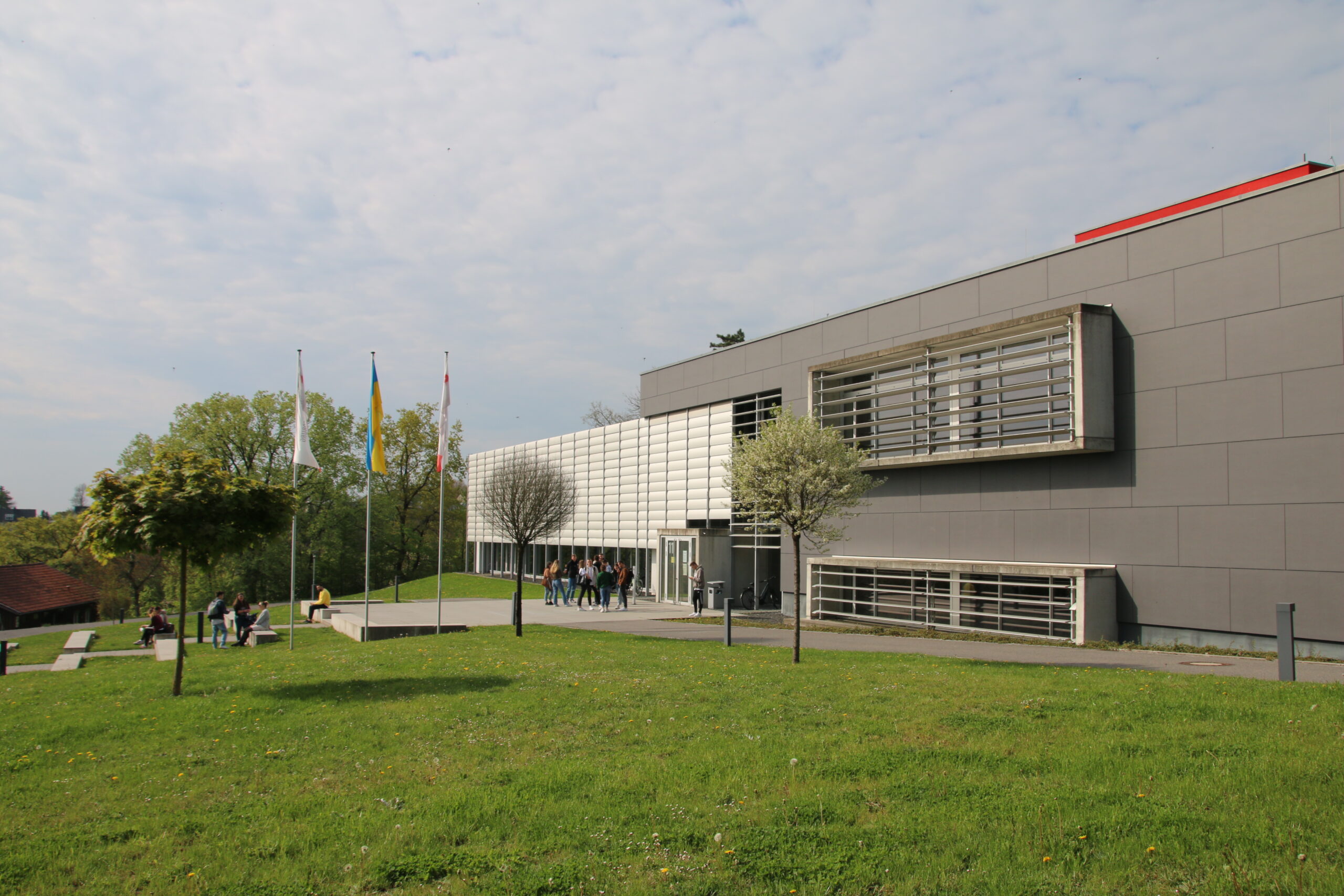 Das moderne Gebäude der Hochschule Coburg zeichnet sich durch eine elegante graue Fassade, große Fenster und einen einzigartigen weißen Bereich aus. Flaggen säumen den Gehweg, während sich Menschen in der Nähe des Eingangs versammeln, während eine Rasenfläche mit Bäumen das Gebäude unter einem teilweise bewölkten Himmel umgibt.