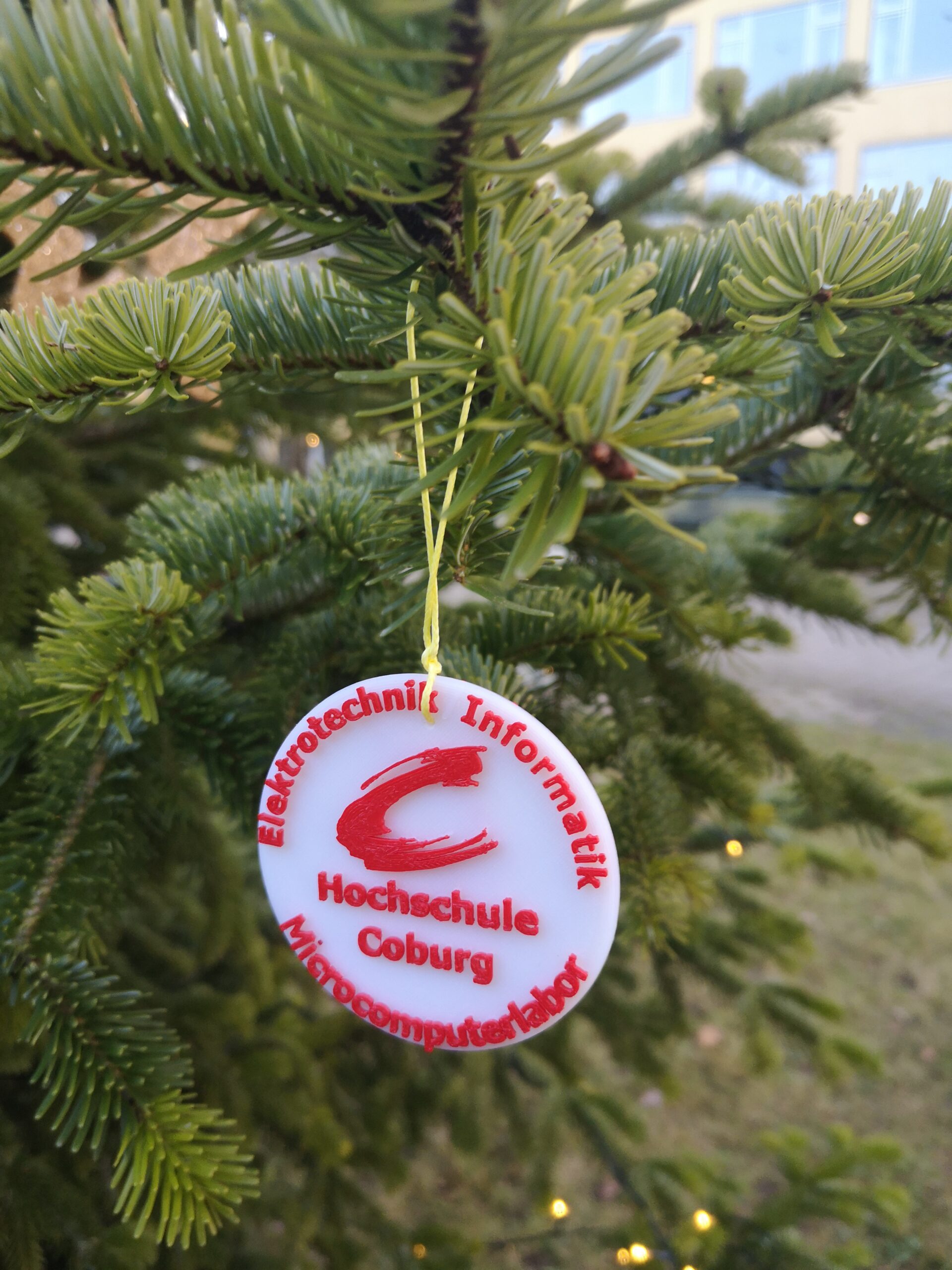 Nahaufnahme eines Weihnachtsbaums, der mit einem runden Ornament geschmückt ist. Dieses Ornament zeigt den leuchtend roten Text: „Elektrotechnik & Informatik, Hochschule Coburg, Minicomputerlabor“ zusammen mit einem stilisierten roten Buchstaben „C“, der den Innovationsgeist der Hochschule Coburg symbolisiert.