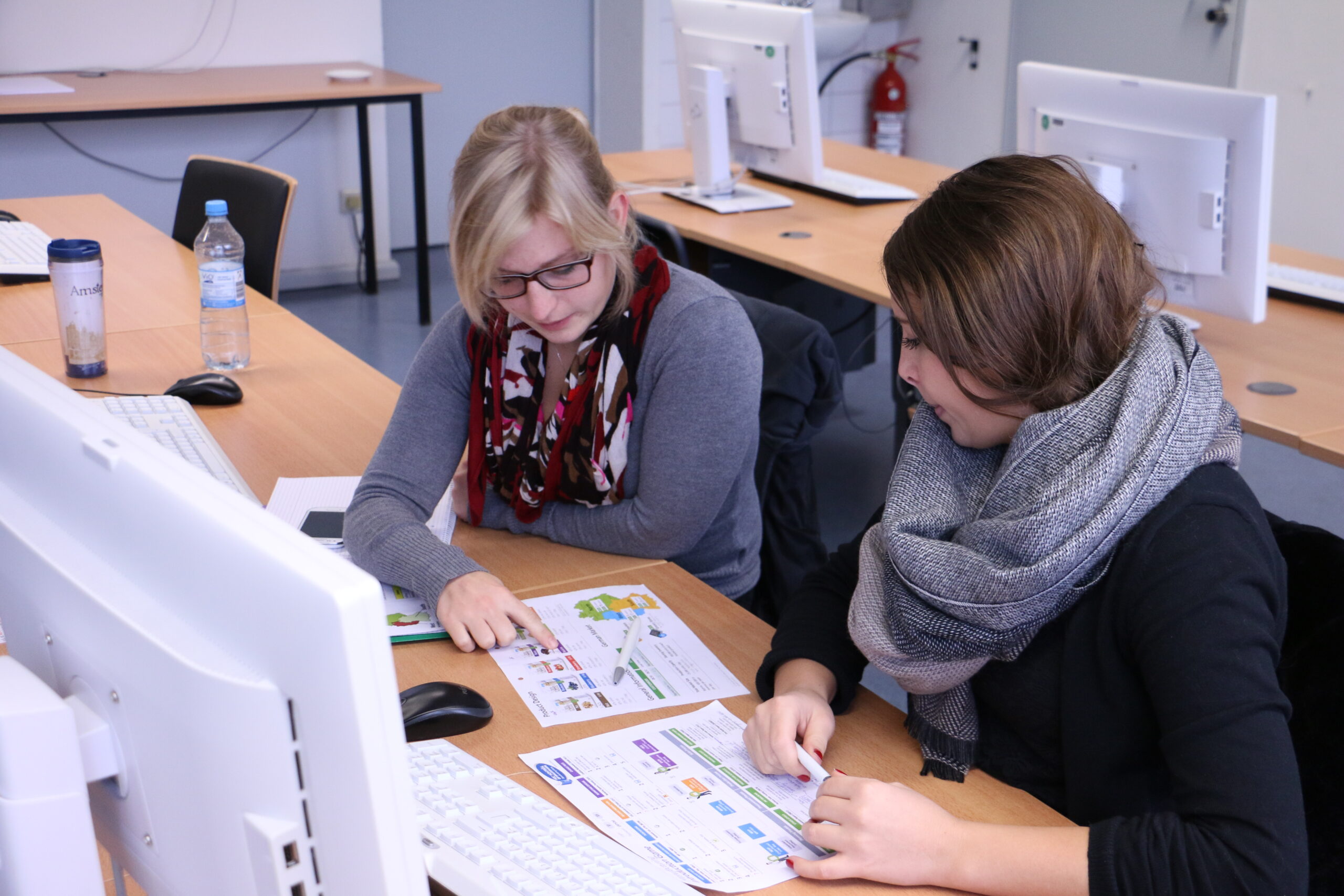 Zwei Personen sitzen an einem Schreibtisch im Computerlabor der Hochschule Coburg und prüfen Drucksachen. Beide tragen Schals und Brillen, umgeben von mehreren Monitoren. Auf dem Tisch stehen eine Flasche Wasser und ein Kaffeebecher.