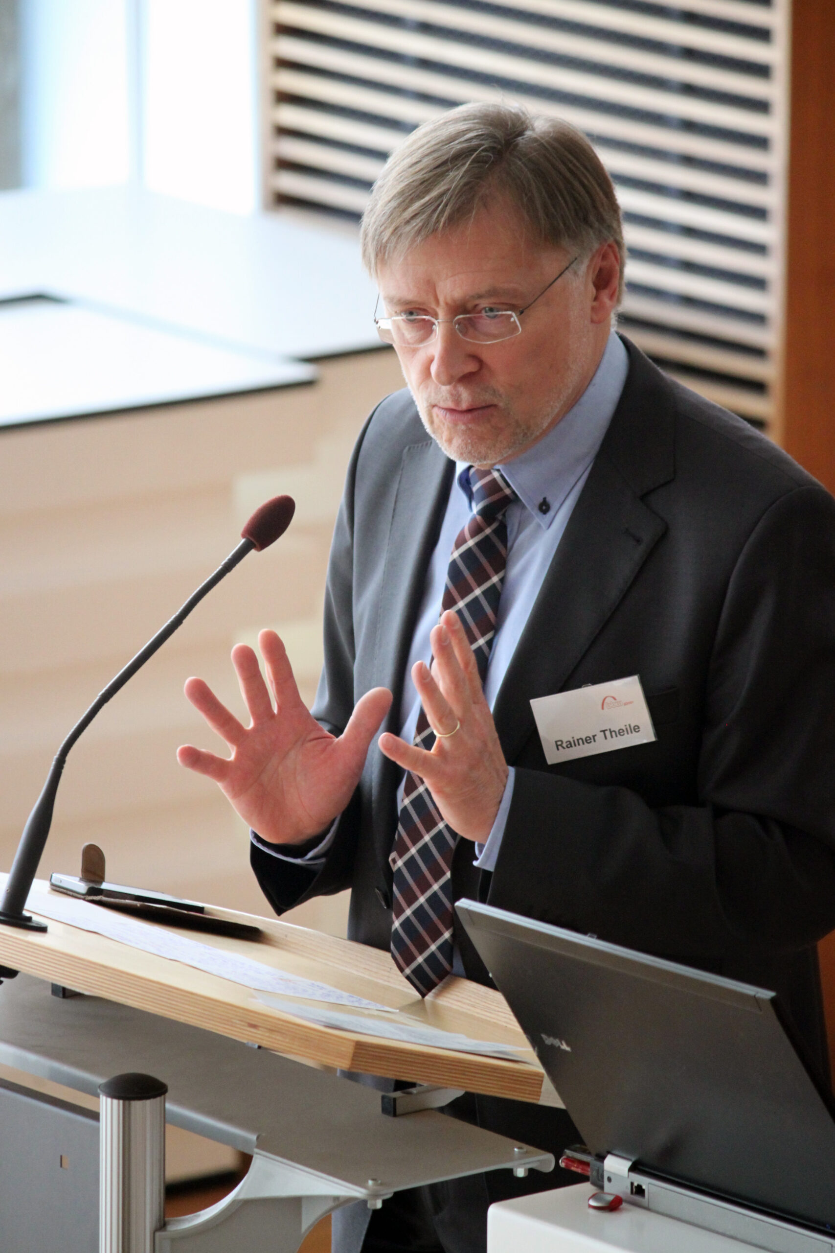 Ein Mann in Anzug und Krawatte, Vertreter der Hochschule Coburg, spricht an einem Podium mit Mikrofon. Er gestikuliert mit offenen Händen und steht vor einem Laptop und Papieren. Ein Namensschild ziert seine Jacke, während er vor einer hellen, gestreiften Wand zum Publikum spricht.