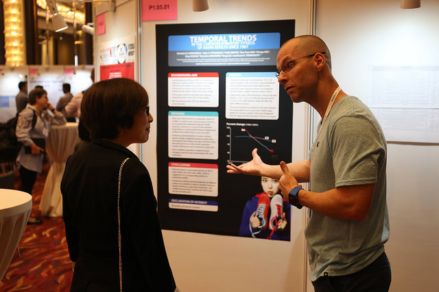 Zwei Personen unterhalten sich vor einem Poster mit dem Titel „Temporale Trends“ in der Ausstellung der Hochschule Coburg. Das Poster enthält Text, Grafiken und Abbildungen. In der Umgebung herrscht reges Treiben mit Konferenzteilnehmern und im Hintergrund sind verschiedene interessante Darstellungen zu sehen.