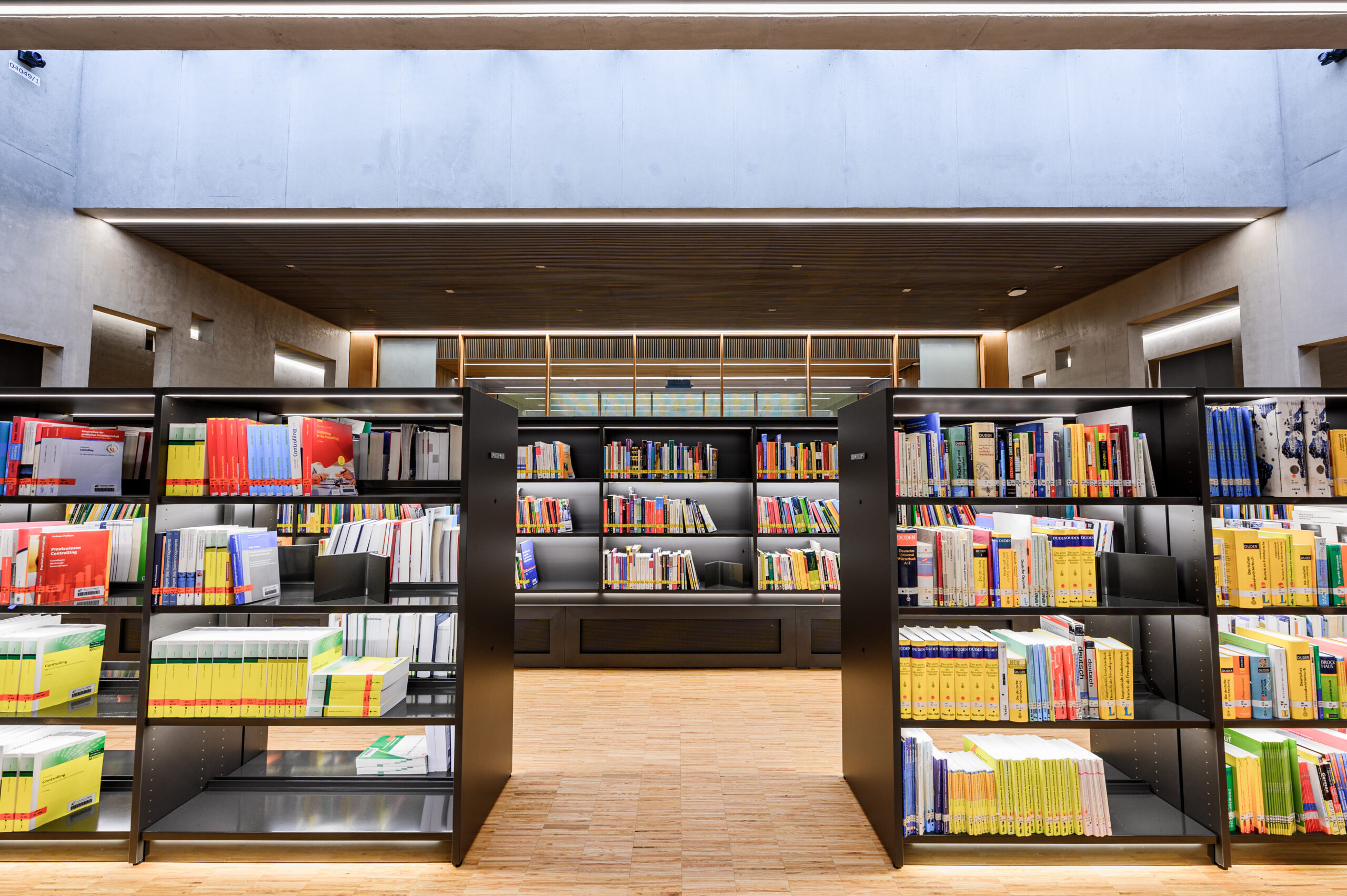 Die moderne Bibliothek der Hochschule Coburg besticht durch zwei Bücherregalreihen voller farbenfroher Bände. Dieser ruhige Raum mit Holzfußboden, hoher Decke und Oberlicht zeichnet sich durch minimalistisches Design aus und schafft eine einladende Atmosphäre zum Lernen und Nachdenken.