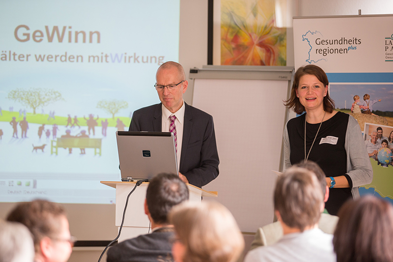 Ein Mann im Anzug und eine Frau im schwarzen Hemd stehen vor einem aufmerksamen Publikum und präsentieren ihre Präsentation. Das Banner der Hochschule Coburg ist deutlich zu sehen. Auf dem Bildschirm im Hintergrund ist Text zu sehen, der zur professionellen Atmosphäre beiträgt.