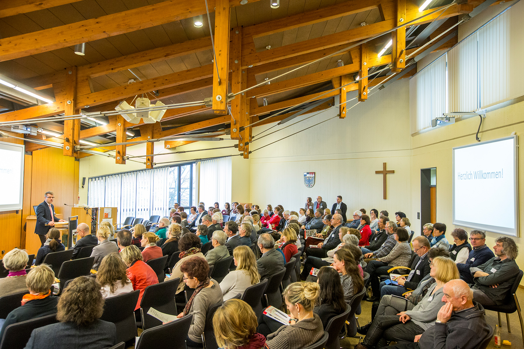 Eine große Gruppe von Menschen sitzt während einer Präsentation an der Hochschule Coburg in einem Raum mit Holzdecke. Links steht ein Redner an einem Podium. Auf einem Bildschirm steht „Herzlich Willkommen!“ auf Deutsch. An der Wand ist ein Holzkreuz angebracht.