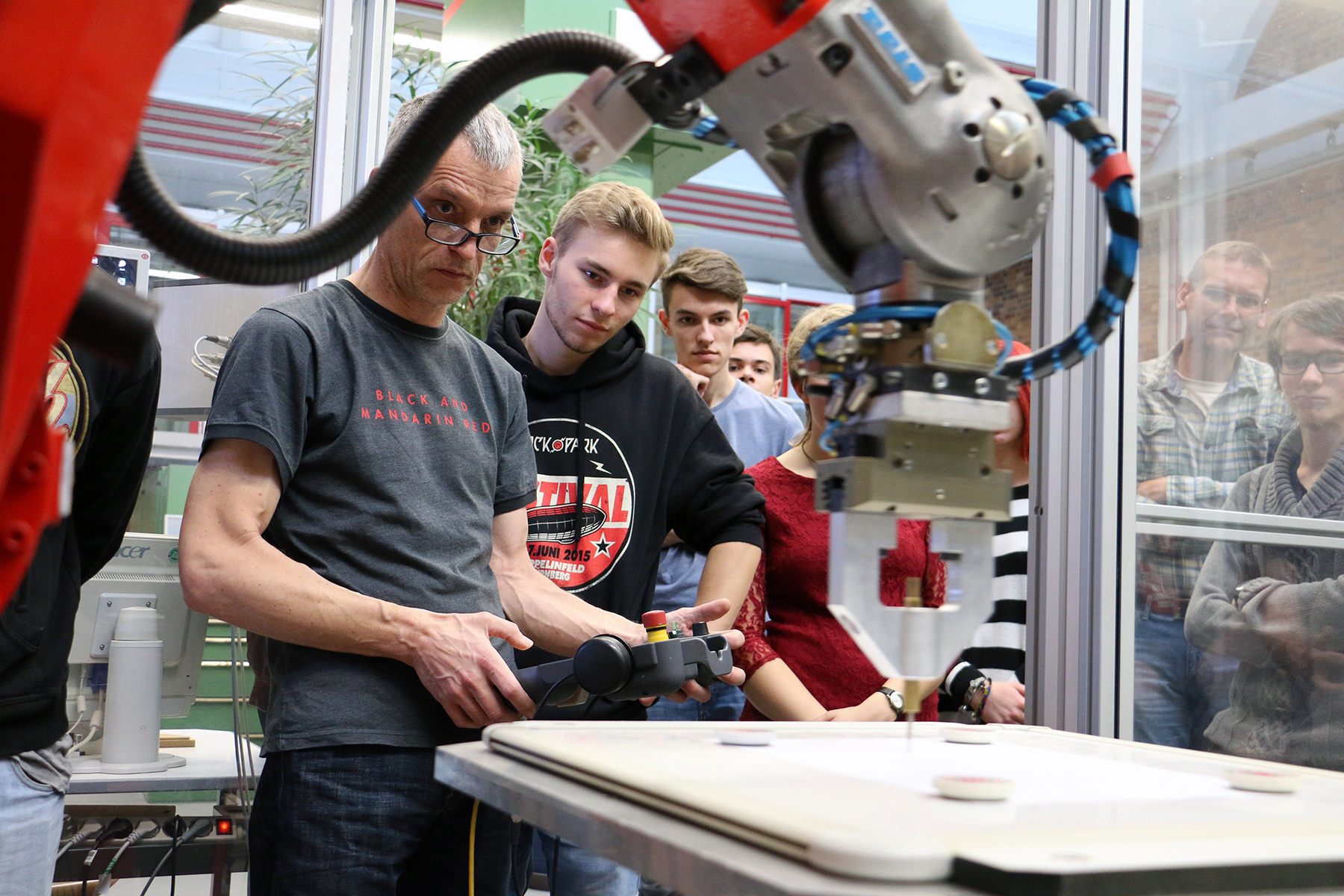 Eine Gruppe von Menschen an der Hochschule Coburg beobachtet einen Mann, der in einer Laborumgebung einen Roboterarm bedient. Der Mann konzentriert sich auf die Verwendung einer Handsteuerung und steuert die Maschine, während sie auf einer flachen Oberfläche arbeitet, während andere aufmerksam zuschauen.