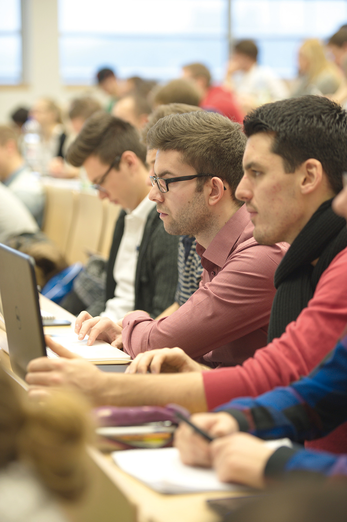 An der Hochschule Coburg machen sich die Studierenden während der Vorlesung aufmerksam Notizen. In Reihen sitzend, mit Laptops und Notebooks, konzentrieren sie sich auf den Vortrag, im Hintergrund fällt Tageslicht durch große Fenster.