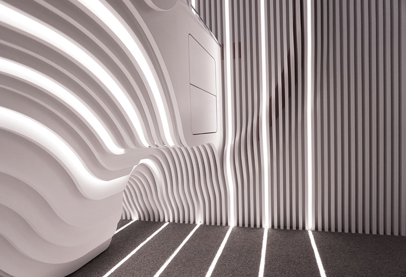 Futuristic interior with wavy white wall panels at Hochschule Coburg, featuring integrated lighting design. The play of light and shadow creates a dynamic and modern atmosphere, while gray flooring complements the sleek aesthetic.