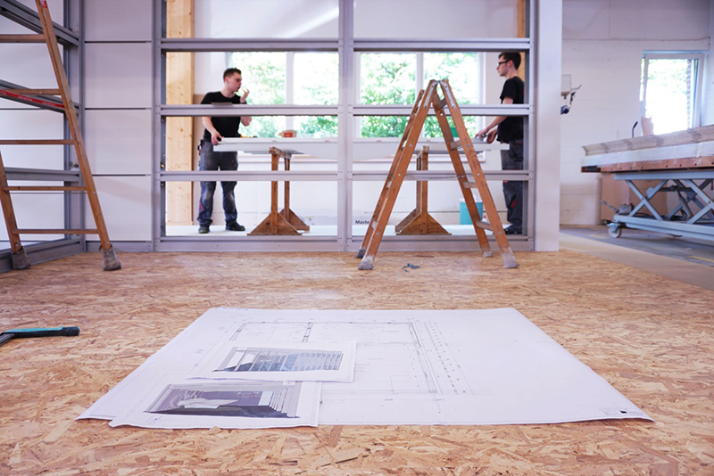 Blueprints are spread out on a wooden floor in front of a metal-frame partition. Two people stand in the background, engaged in discussion near a wooden table and ladders, with views of greenery outside—a typical scene at Hochschule Coburg's architectural design class.