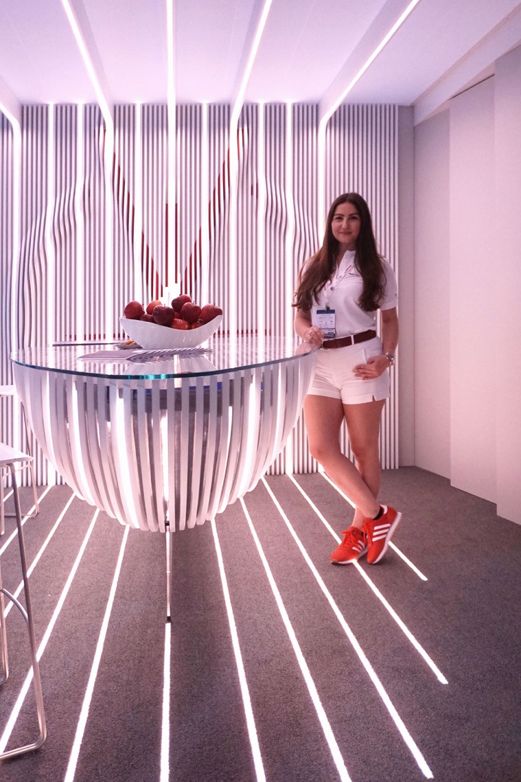 A woman in a white outfit and red sneakers stands next to a modern, round glass table with vertical stripes. The room, inspired by Hochschule Coburg's design principles, features striped lighting patterns on the floor and walls, with a bowl of red apples on the table.