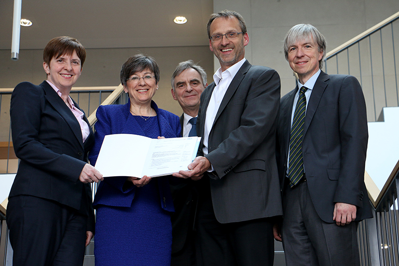 Eine Gruppe von fünf Personen, drei Männer und zwei Frauen, steht auf einer Treppe im Innenbereich der Hochschule Coburg. Sie lächeln und halten gemeinsam ein Dokument in der Hand. Sie strahlen eine professionelle und formelle Ausstrahlung aus.