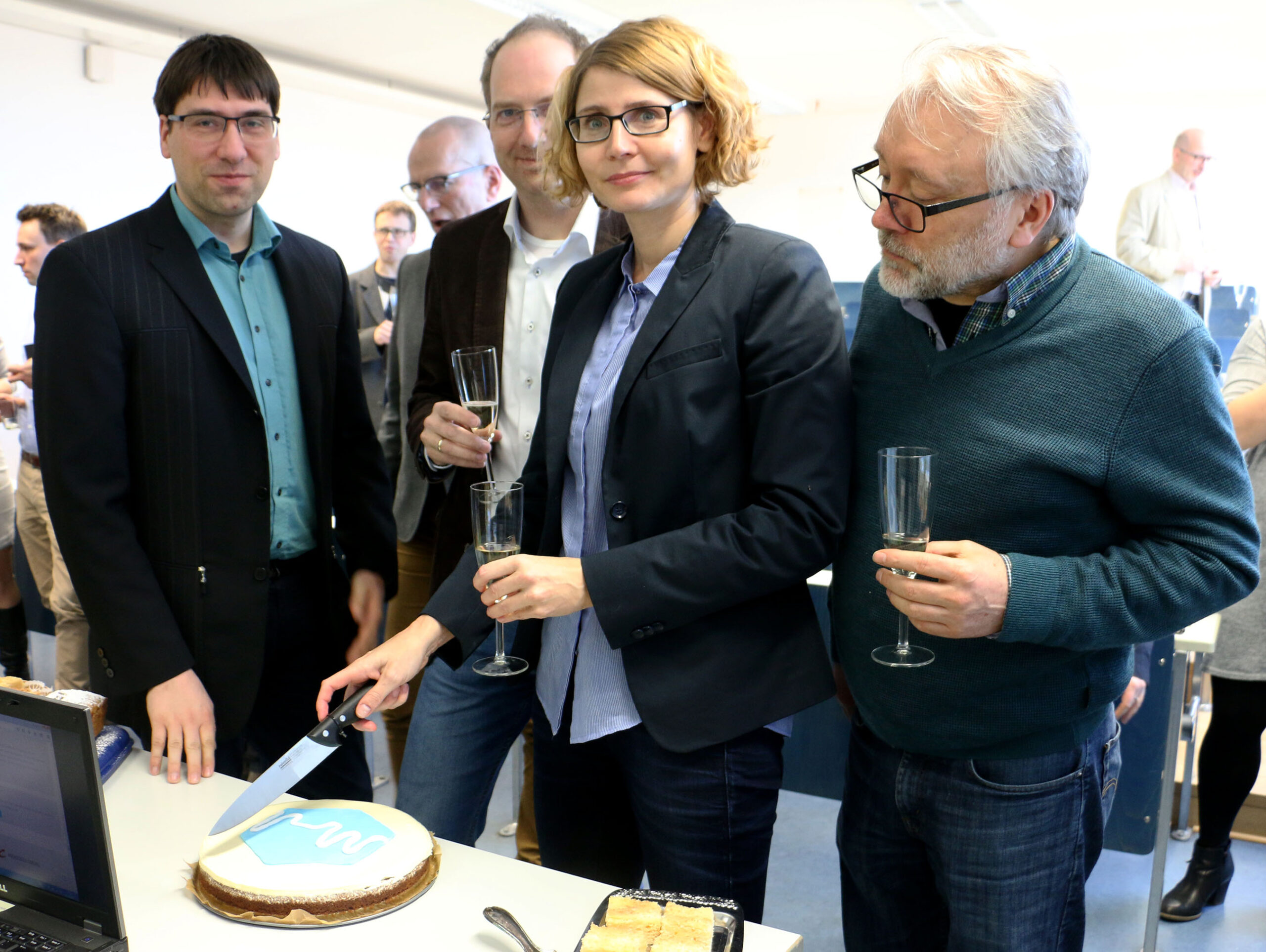 Bei einer Veranstaltung an der Hochschule Coburg schneidet eine Frau eine mit einem blau-weißen Muster verzierte Torte an. Neben ihr erheben drei Männer ihre Sektgläser, während andere im Hintergrund miteinander plaudern und für eine festliche Atmosphäre sorgen.