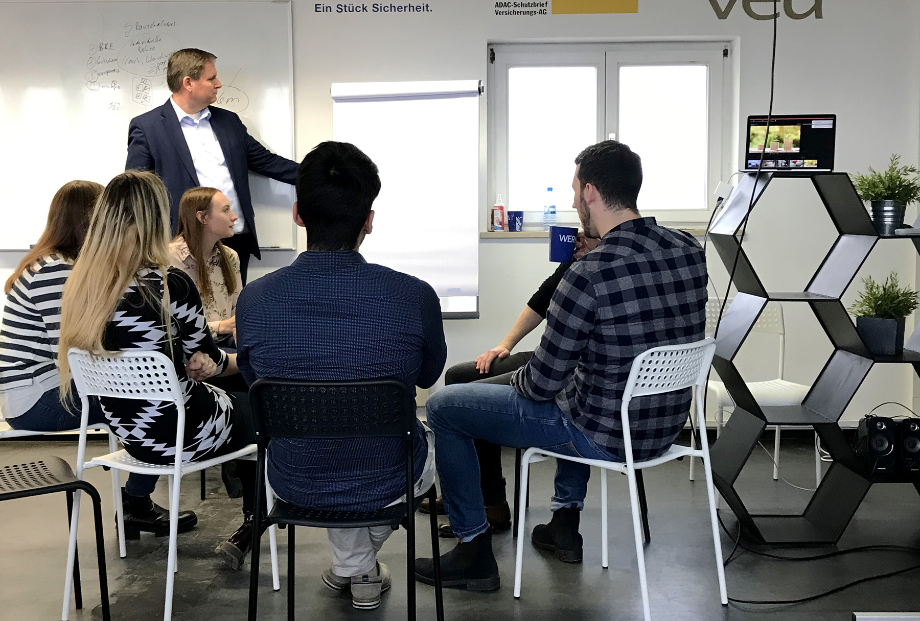 Ein Mann steht an einem Flipchart und präsentiert vor sechs aufmerksamen Teilnehmern in einem Raum, der von der modernen Designästhetik der Hochschule Coburg inspiriert ist und über sechseckige Regale und ein großes Fenster verfügt. Ein Fernseher zeigt mehrere Bilder und verstärkt so die konzentrierte Atmosphäre.