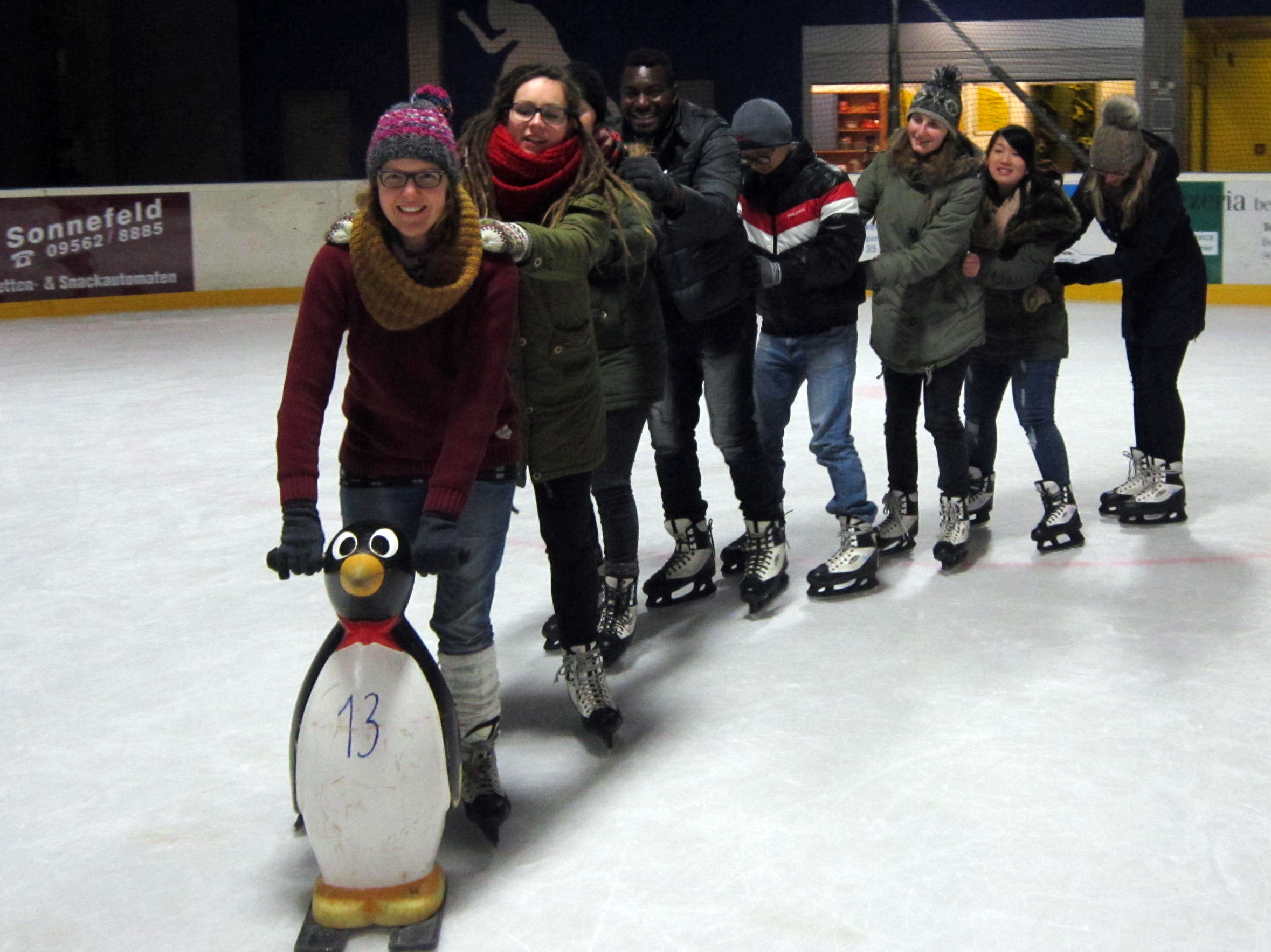 Eine Reihe von Menschen läuft in der Halle der Hochschule Coburg Schlittschuh. Sie halten sich aneinander fest und an der Spitze steht eine Pinguin-Schlittschuhhilfe. Sie lächeln, tragen Winterkleidung und scheinen Spaß in dieser lebendigen Universitätsumgebung zu haben.