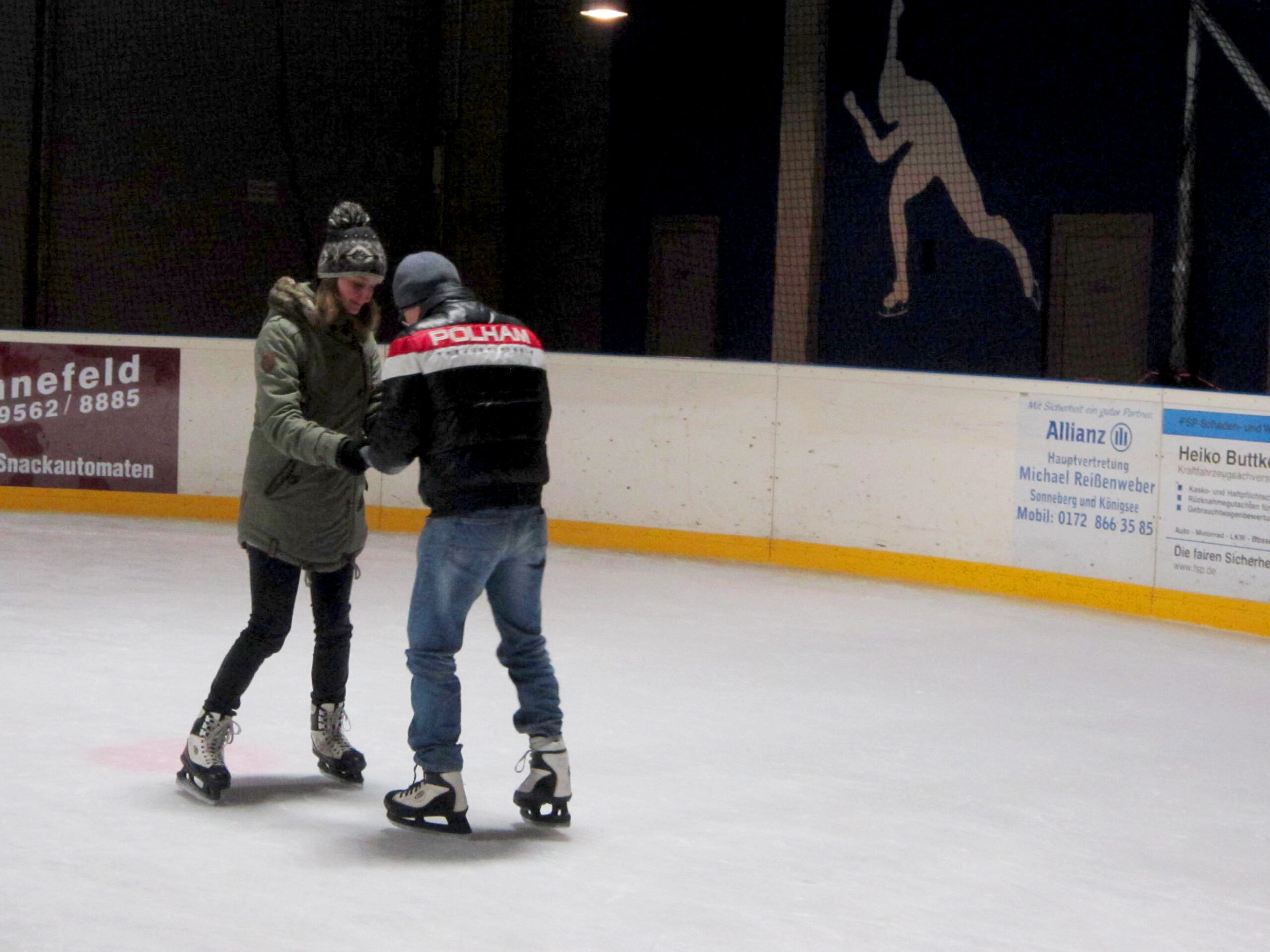 Bei einer Veranstaltung der Hochschule Coburg gleiten zwei Menschen anmutig über die Eisbahn. Einer, in grüner Jacke und schwarzer Hose, hält Händchen mit seinem Partner in schwarz-roter Jacke und Jeans. Die elegante Silhouette eines Schlittschuhläufers ziert die Wand im Hintergrund und fängt die Essenz winterlicher Romantik ein.