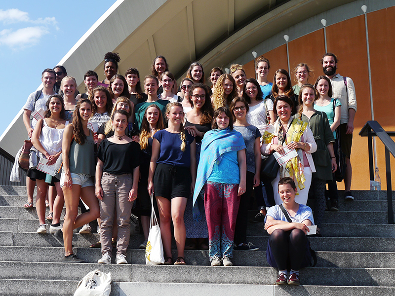 Eine große Gruppe leger gekleideter und lächelnder Menschen versammelt sich auf den sonnenbeschienenen Stufen vor einem modernen Gebäude. Einige sitzen, andere stehen und halten einen Moment an der Hochschule Coburg fest.